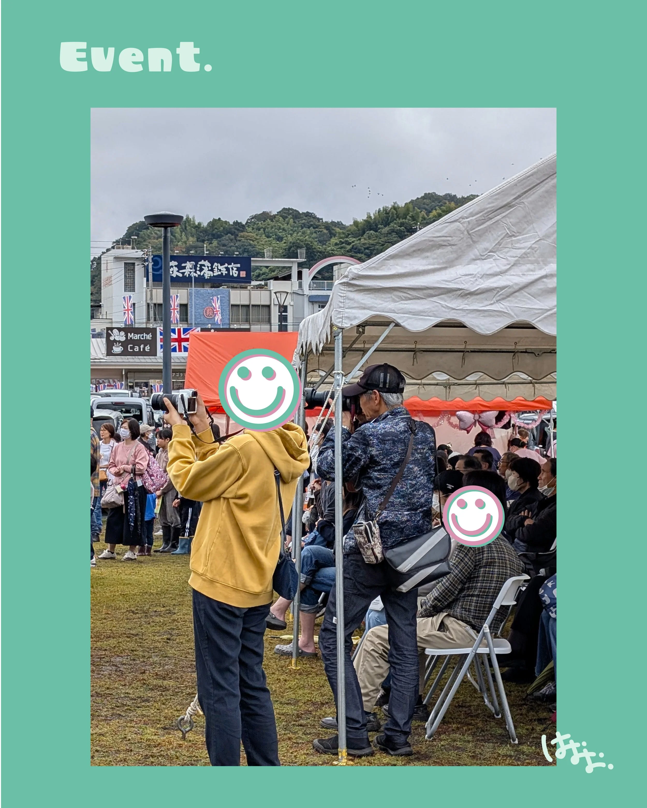 屋外のイベント会場で、多くの人が座っている。左側に黄色いジャケットを着た人がスマートフォンで写真を撮っている。背景には建物や山、空模様が見える。