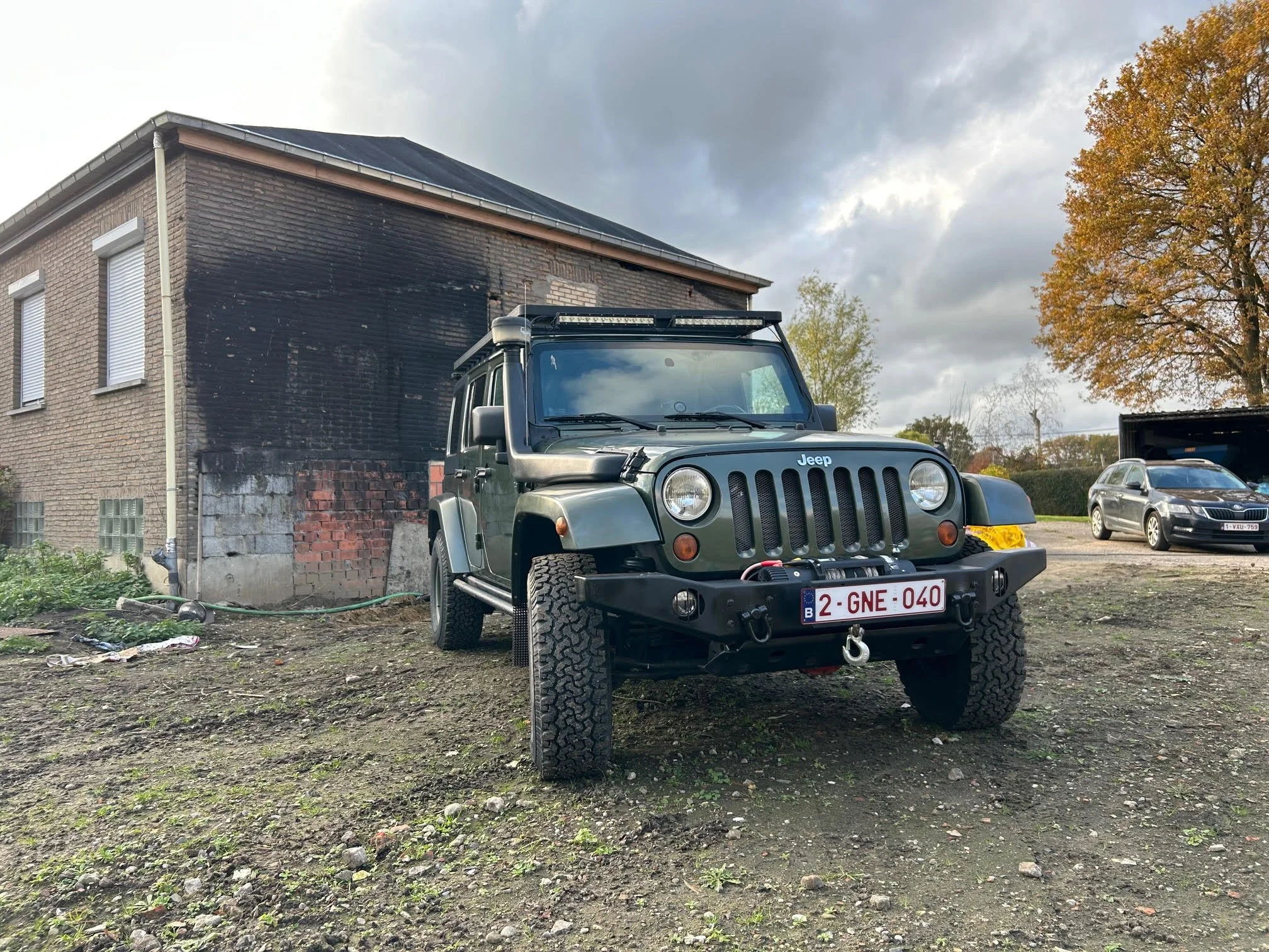 A Jeep off-road vehicle parked outdoors on uneven dirt ground in front of a brick building with black smudges on the wall. There is a white license plate with red and blue details. Two other cars are visible in the background, and trees with autumn foliage are seen to the right.