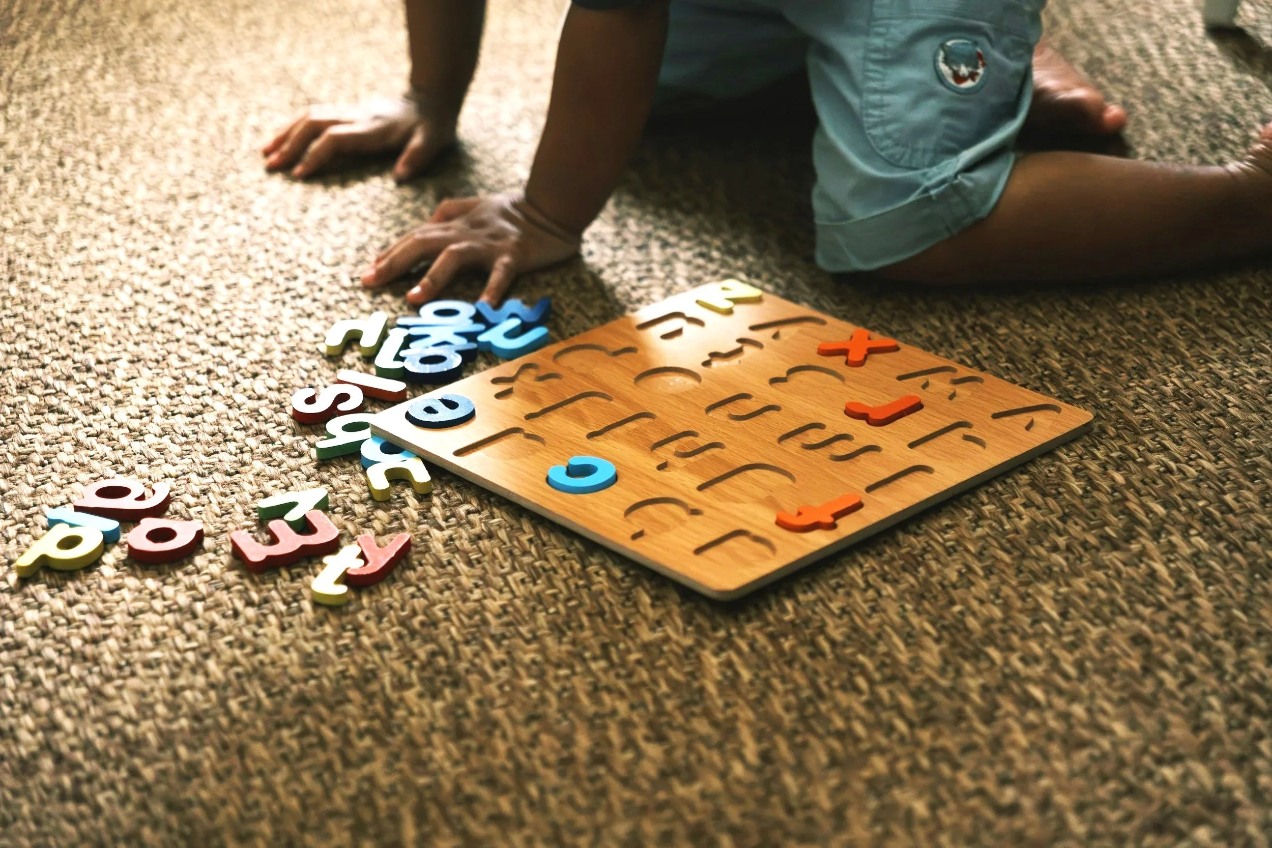 A child playing with colorful alphabet puzzle pieces on a textured carpet. The child is kneeling on the carpet, and the puzzle board is placed in front of them.