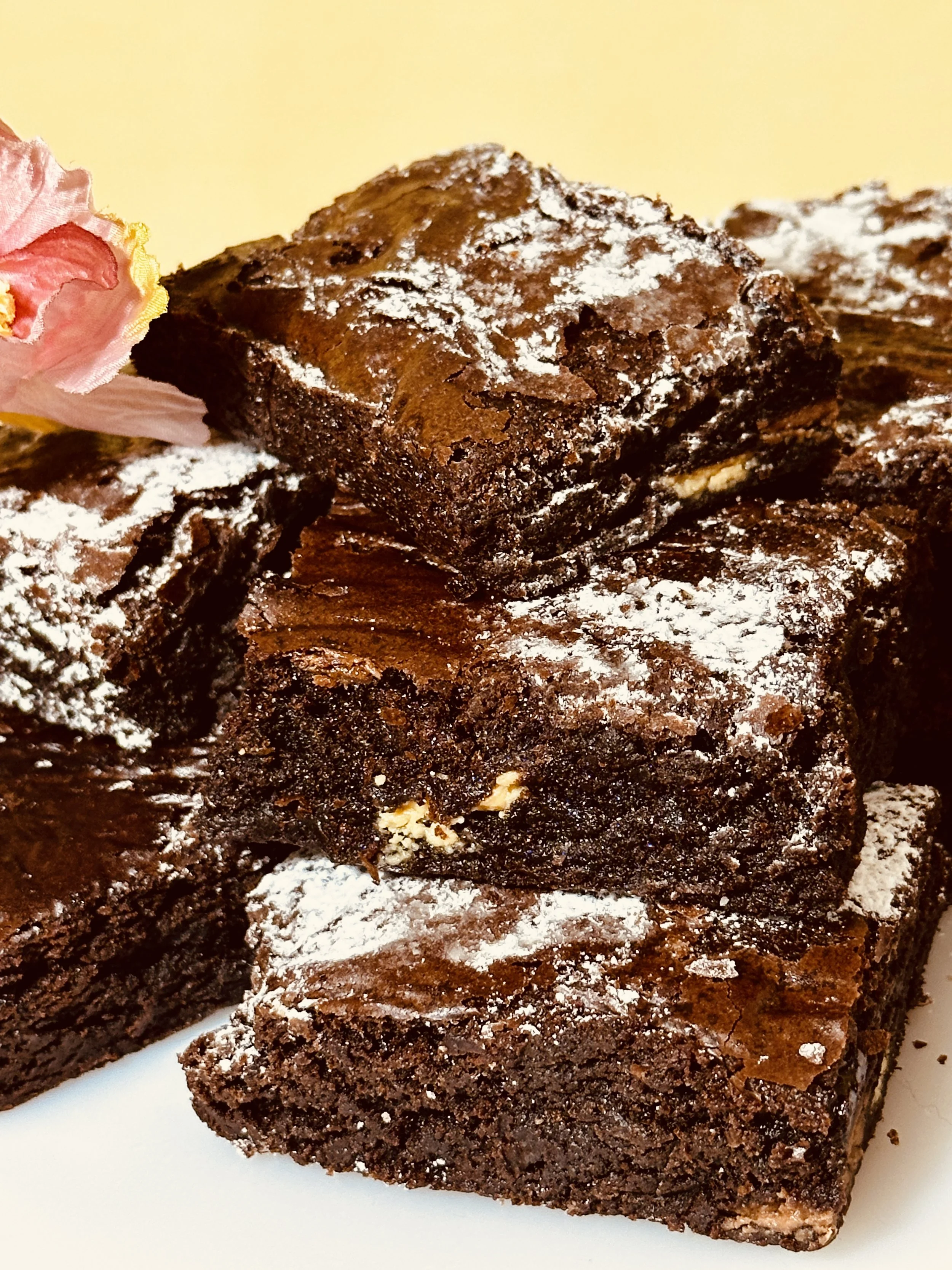 Stack of four chocolate brownies sprinkled with powdered sugar, with a pink flower on the side.