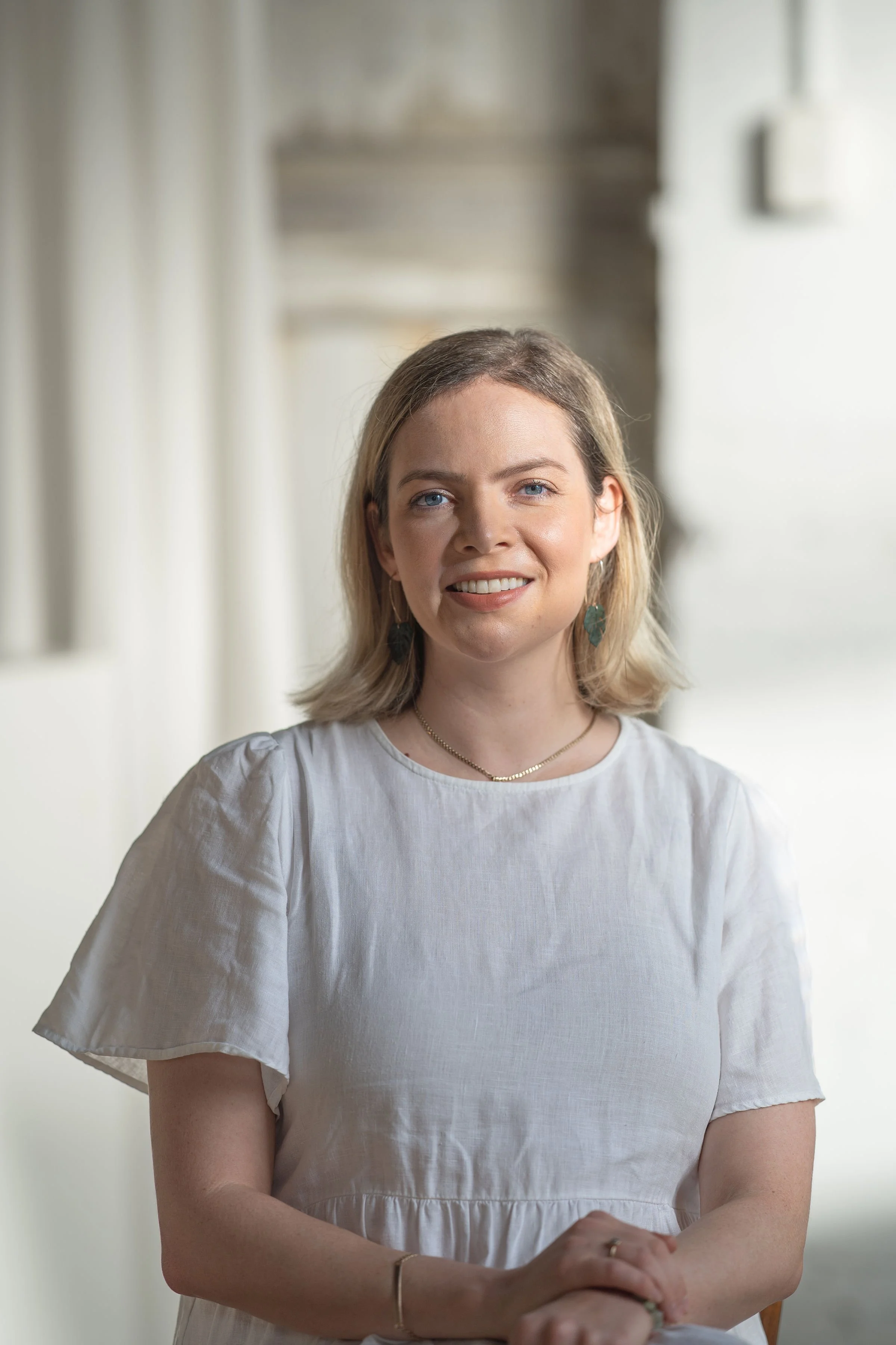 A woman with shoulder-length blonde hair, wearing a white dress, jewelry, and earrings, smiling in a well-lit indoor setting.