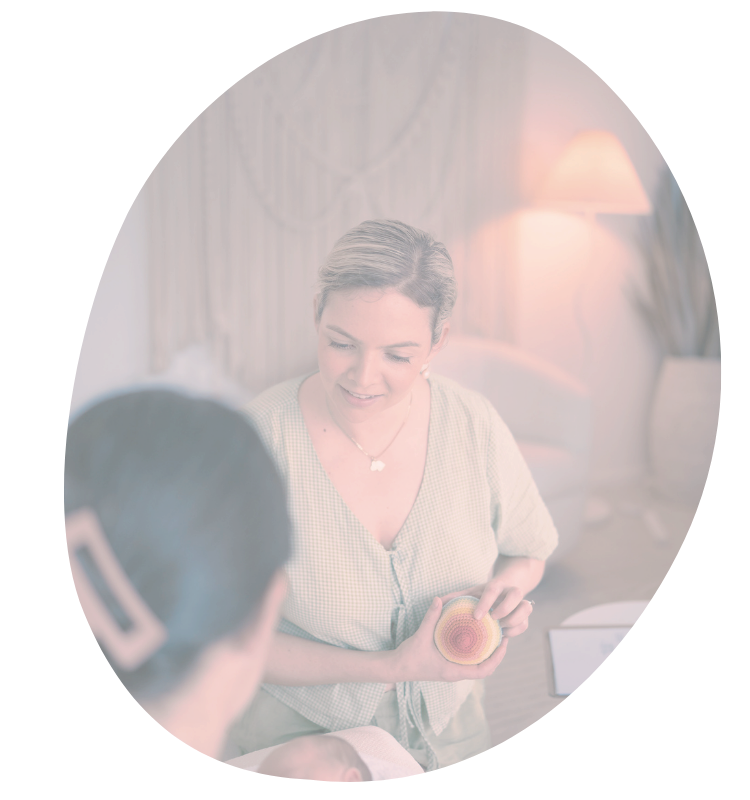 A woman holding a model of a human cell, talking with another woman with glasses in a cozy, softly lit room.