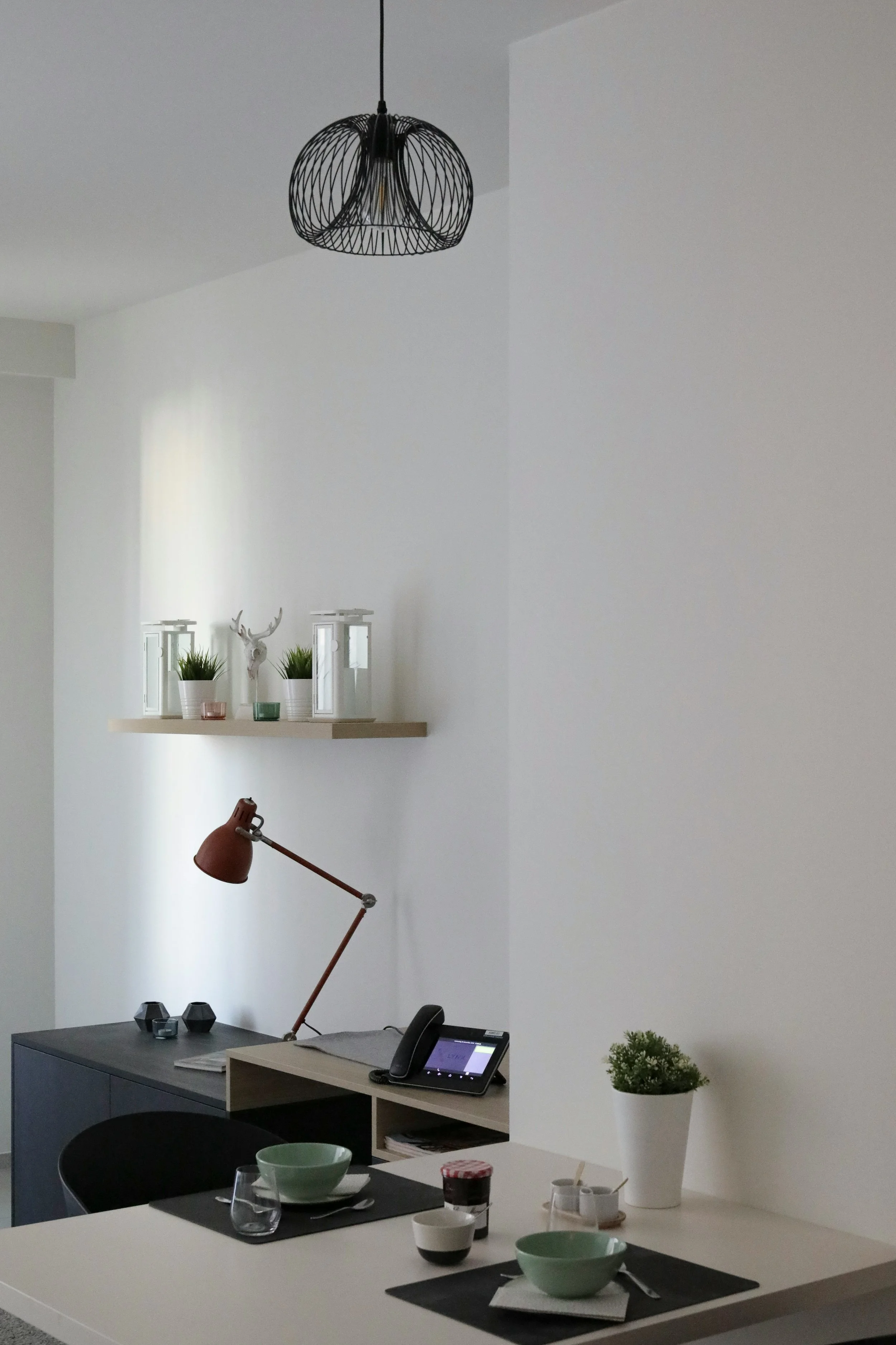 Minimalist interior with a black glass dining table, three black chairs, one gray chair, and decorative white vases with labels on a tray, placed against a textured beige wall with vertical fabric panels.