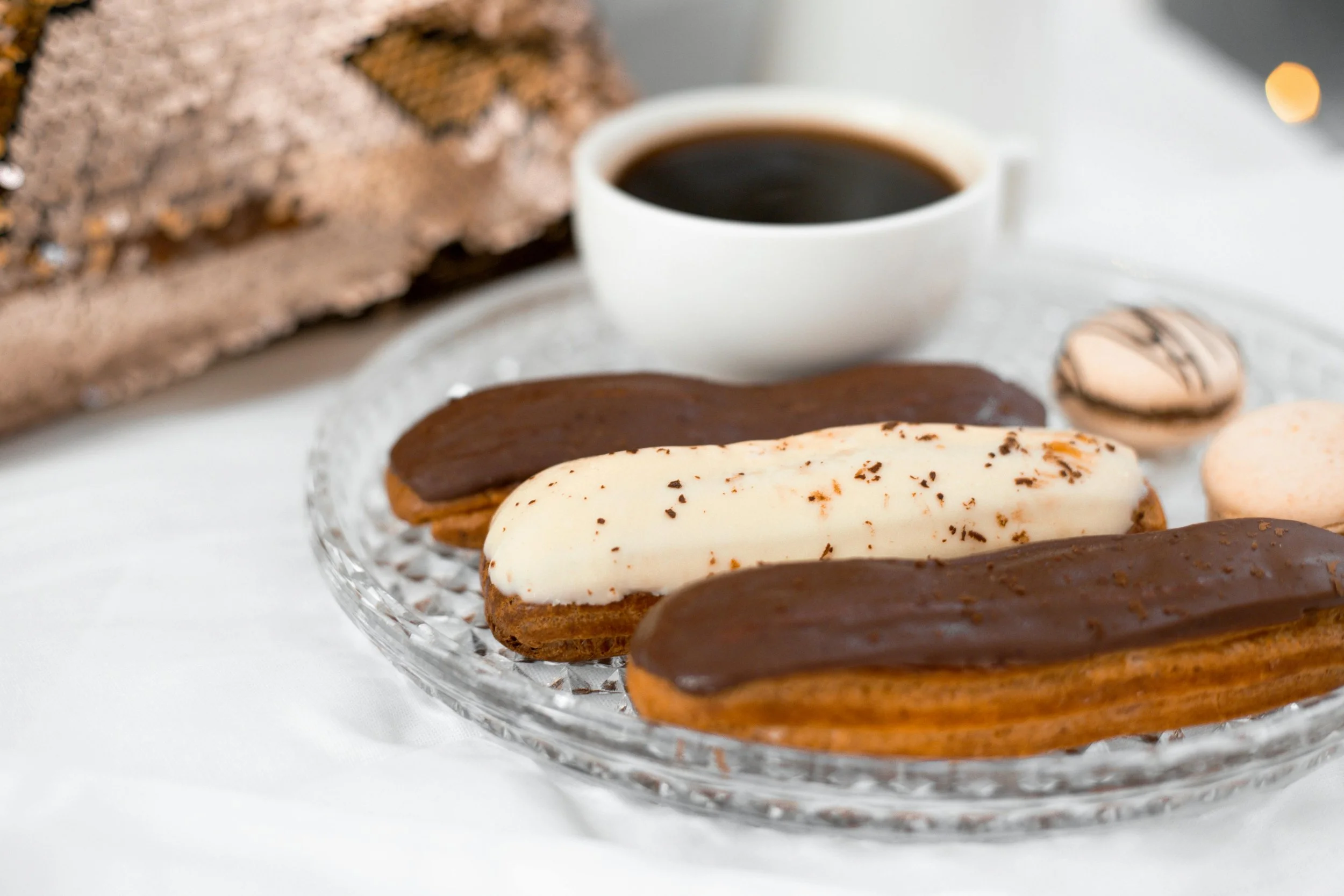 Assorted éclairs, a cup of black coffee, and macarons on a glass platter.