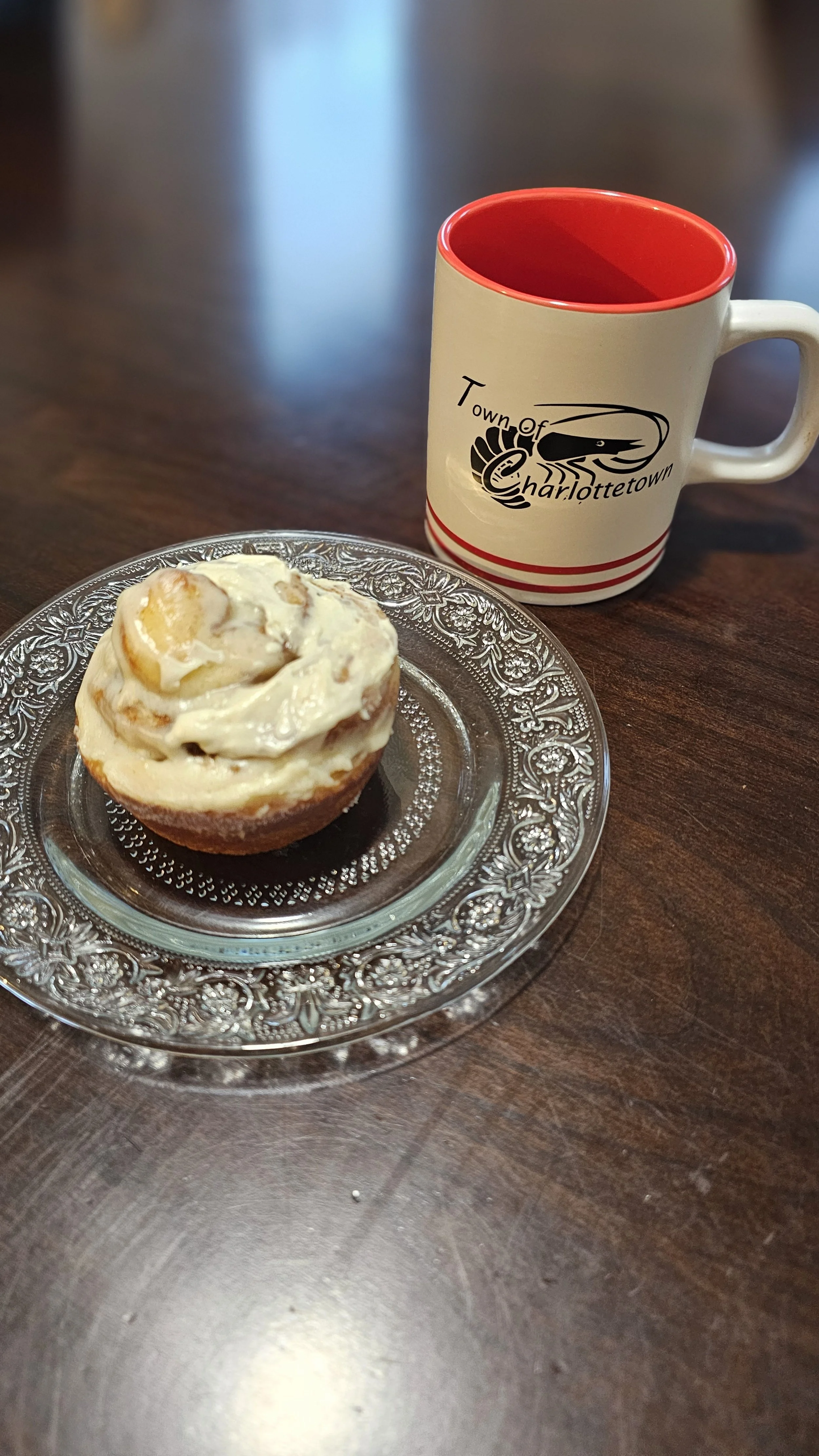 A cupcake with white frosting on a clear glass plate next to a white mug with red interior and the Town of Charlottetown logo, on a dark wooden table.