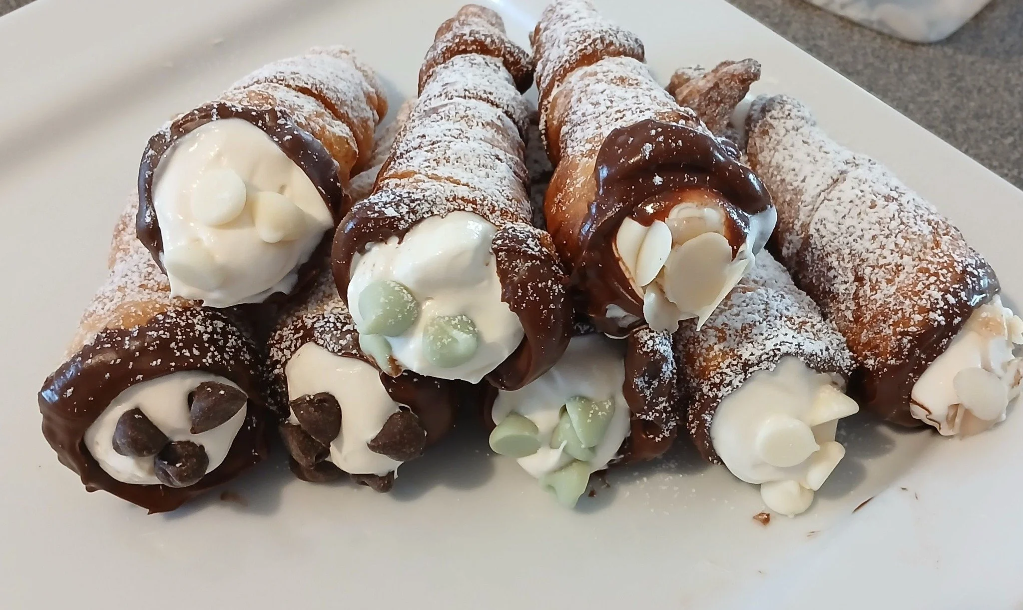 Assorted cannoli on a white plate with chocolate and white chocolate decorations, powdered sugar, and cream filling.