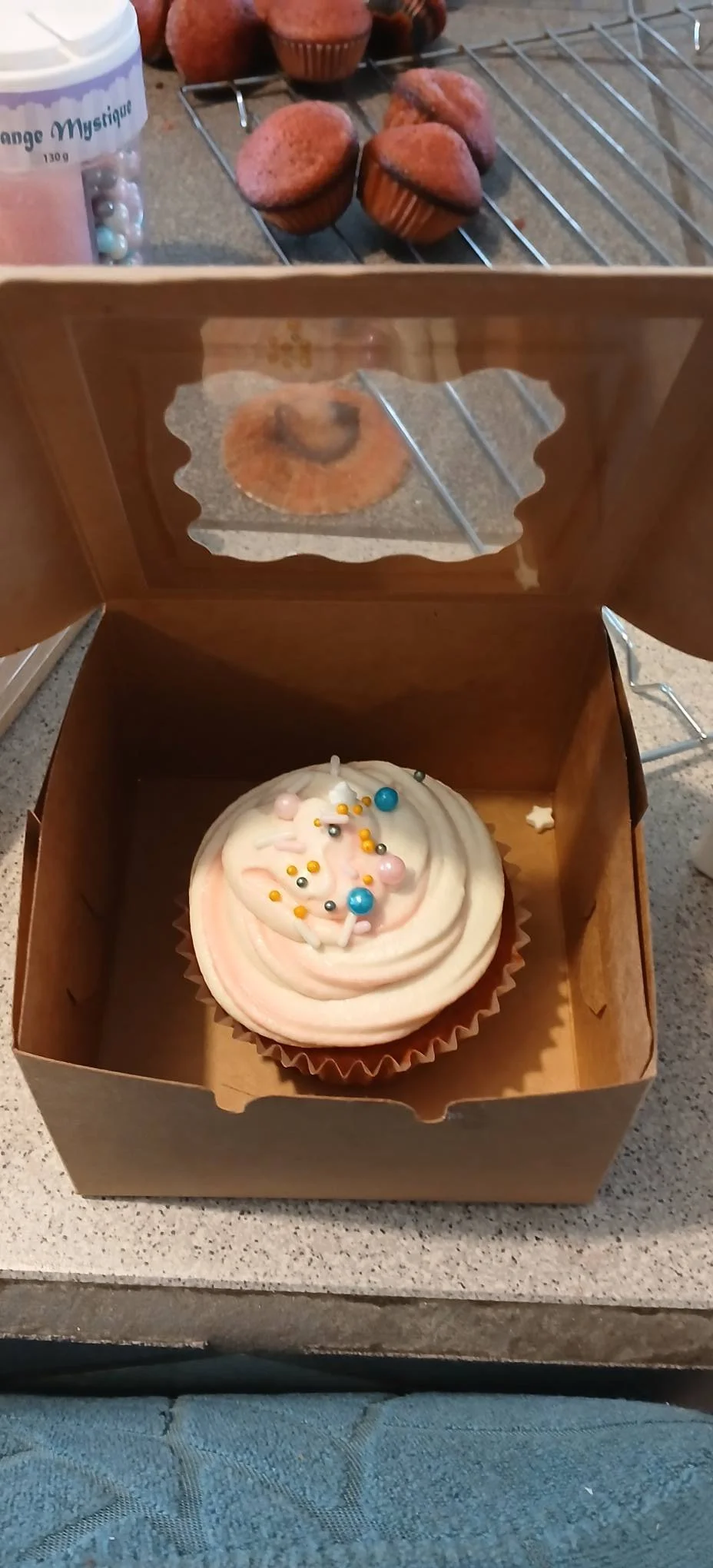 A cupcake with pastel pink and white swirled frosting topped with colorful sprinkles in a baker's box. In the background, a cooling rack holds several plain muffins, and a container of colored pearls is partially visible.