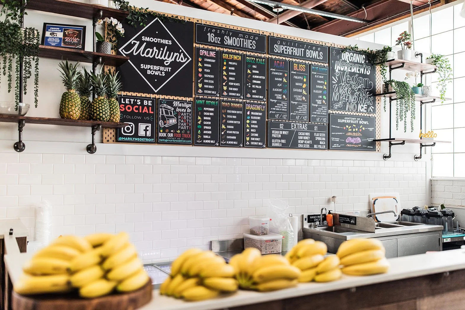 Interior of a smoothie shop with a black chalkboard menu featuring various superfruit bowls and smoothies. Fresh pineapples are displayed on a wooden tray in the foreground, and the wall has white subway tiles with shelves decorated with plants and signs.