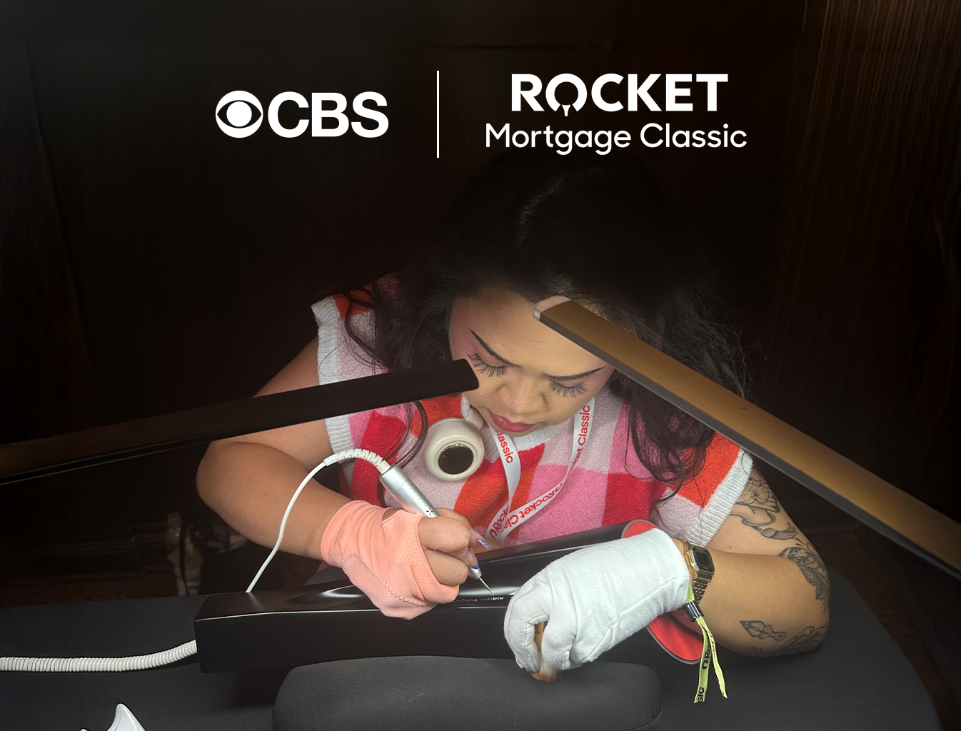 A woman at a desk writing with a pen, wearing a pink and red striped shirt and a white glove, with a CBS Rocket Mortgage Classic logo overhead.