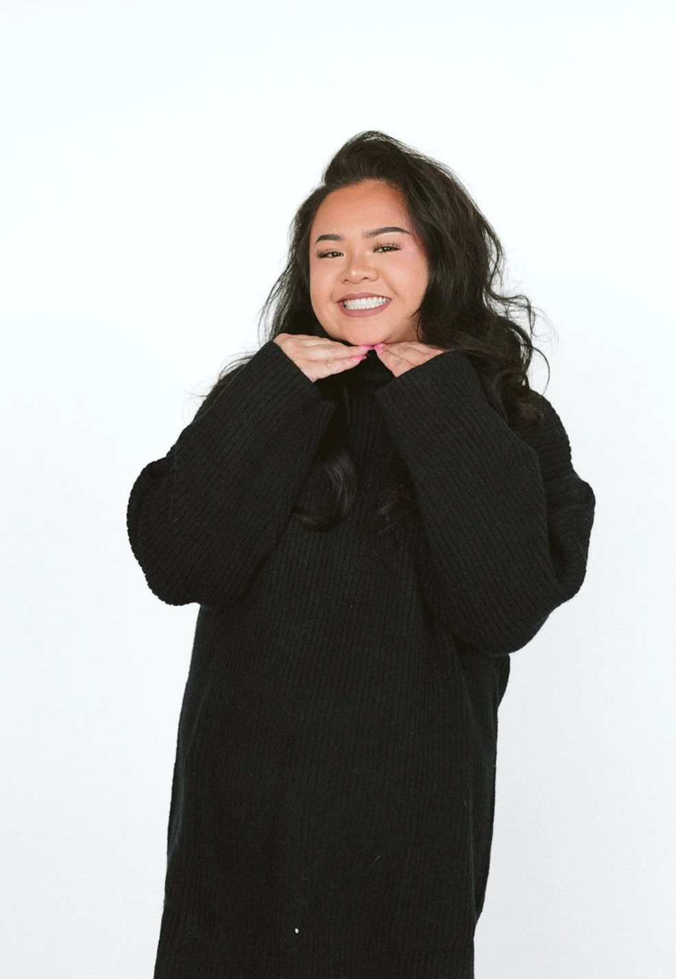 Smiling woman with long dark hair in a black sweater, posing with her hands under her chin, against a plain white background.