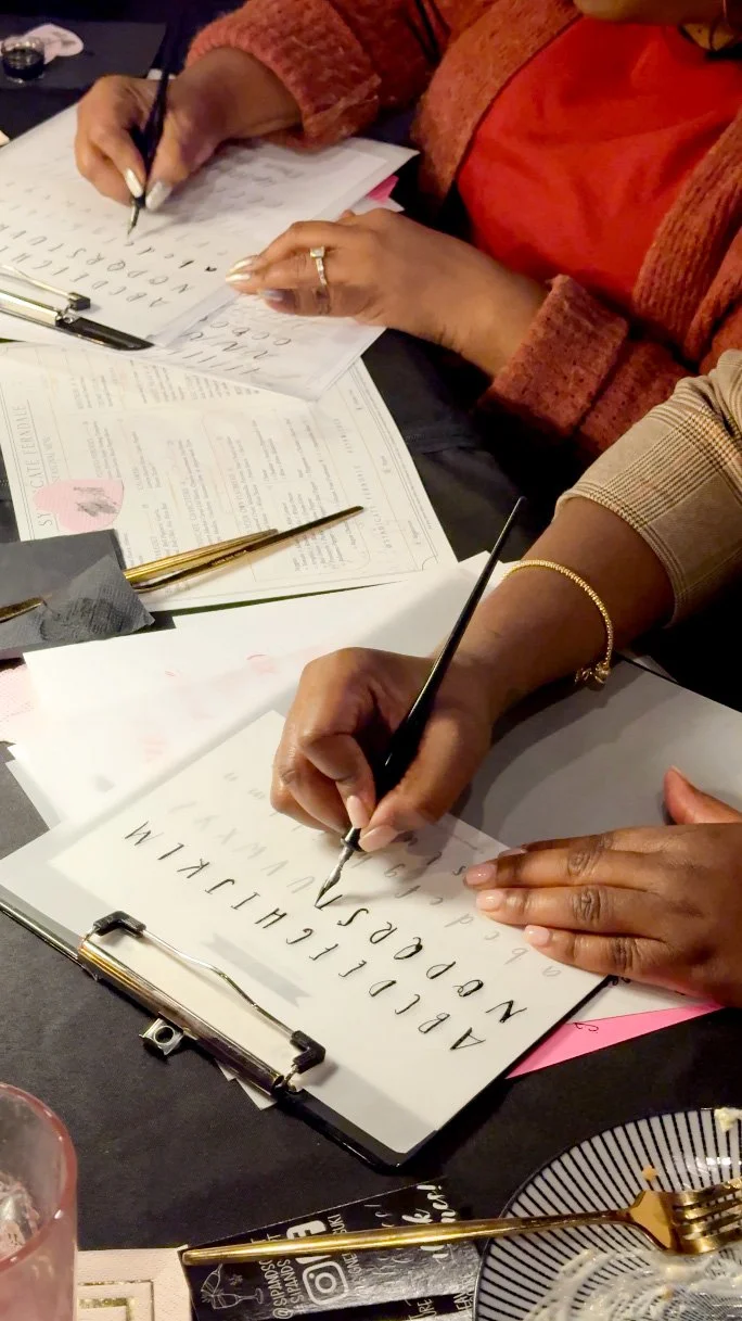 People practicing calligraphy at a table, focusing on writing the alphabet with black ink pens on white paper.