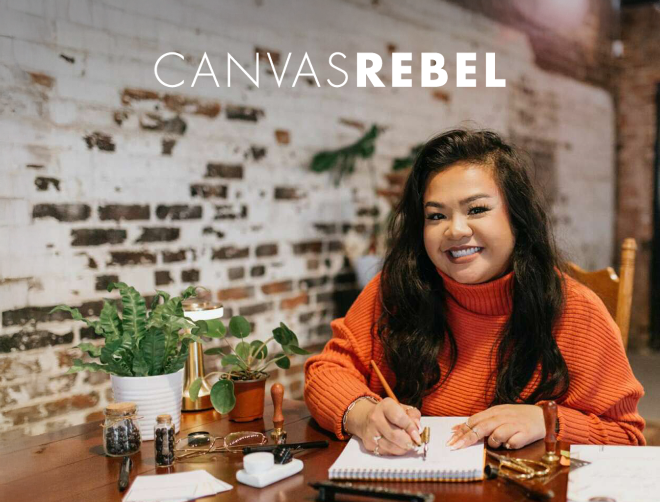 A woman with dark wavy hair and a bright orange sweater, smiling while working at a desk with office supplies, plants, and a brick wall background.