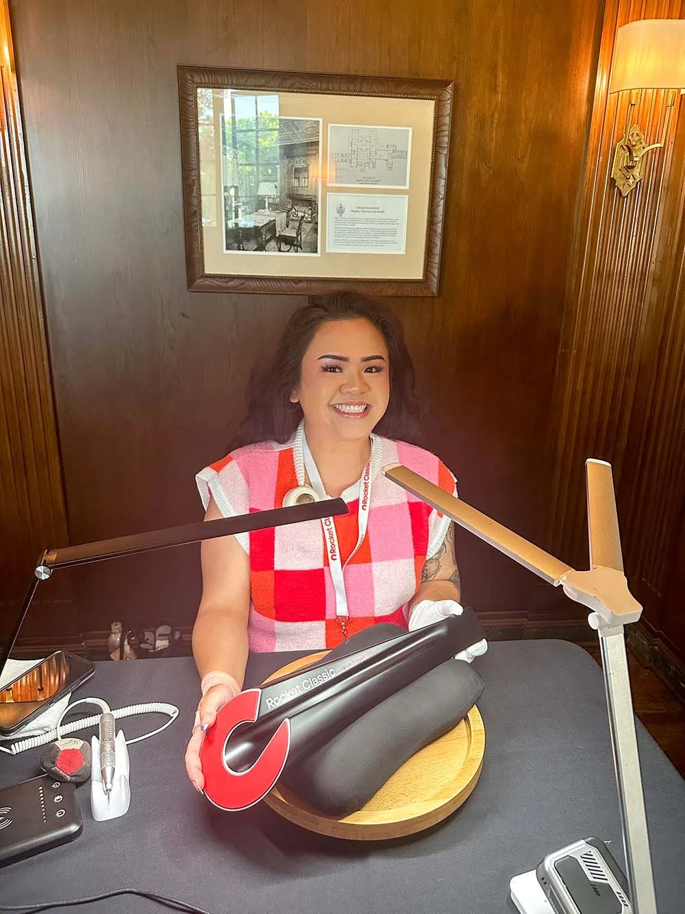 A woman with curly brown hair, smiling, sitting at a desk with a large, red and black microphone labeled 'Rocket Classic' in front of her. She is wearing a sleeveless red, pink, and white checkered top and a lanyard. There are desk lamps, a phone, and various equipment on the desk. The background features wooden wall paneling with a framed picture or document behind her.