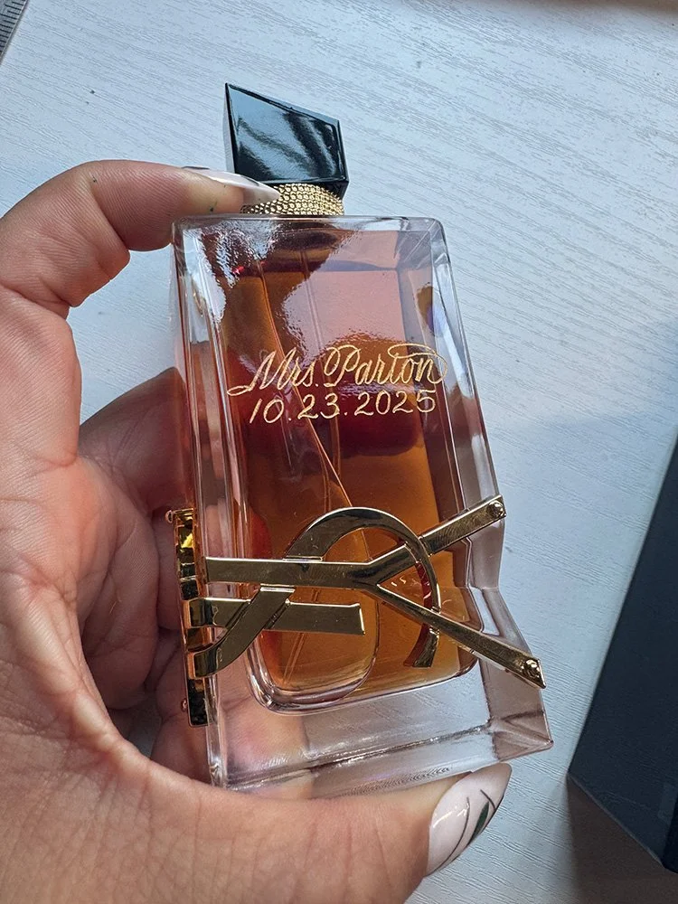 A person holding a rectangular glass perfume bottle with gold accents, engraved with "Mrs. Parton 10.23.2025" on the front, with a black and gold cap on top, placed on a light-colored wooden surface.