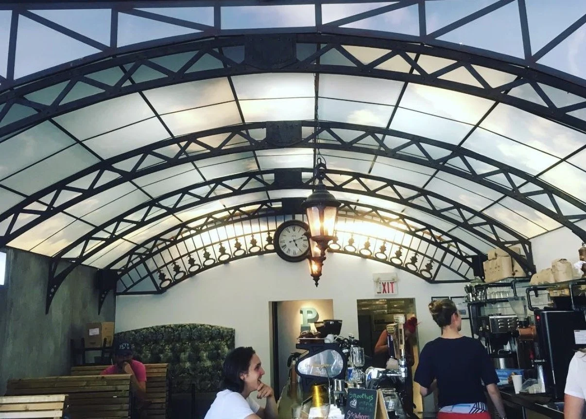 Interior of a coffee shop with a curved glass and metal ceiling, a clock, hanging lantern light, and baristas preparing drinks.