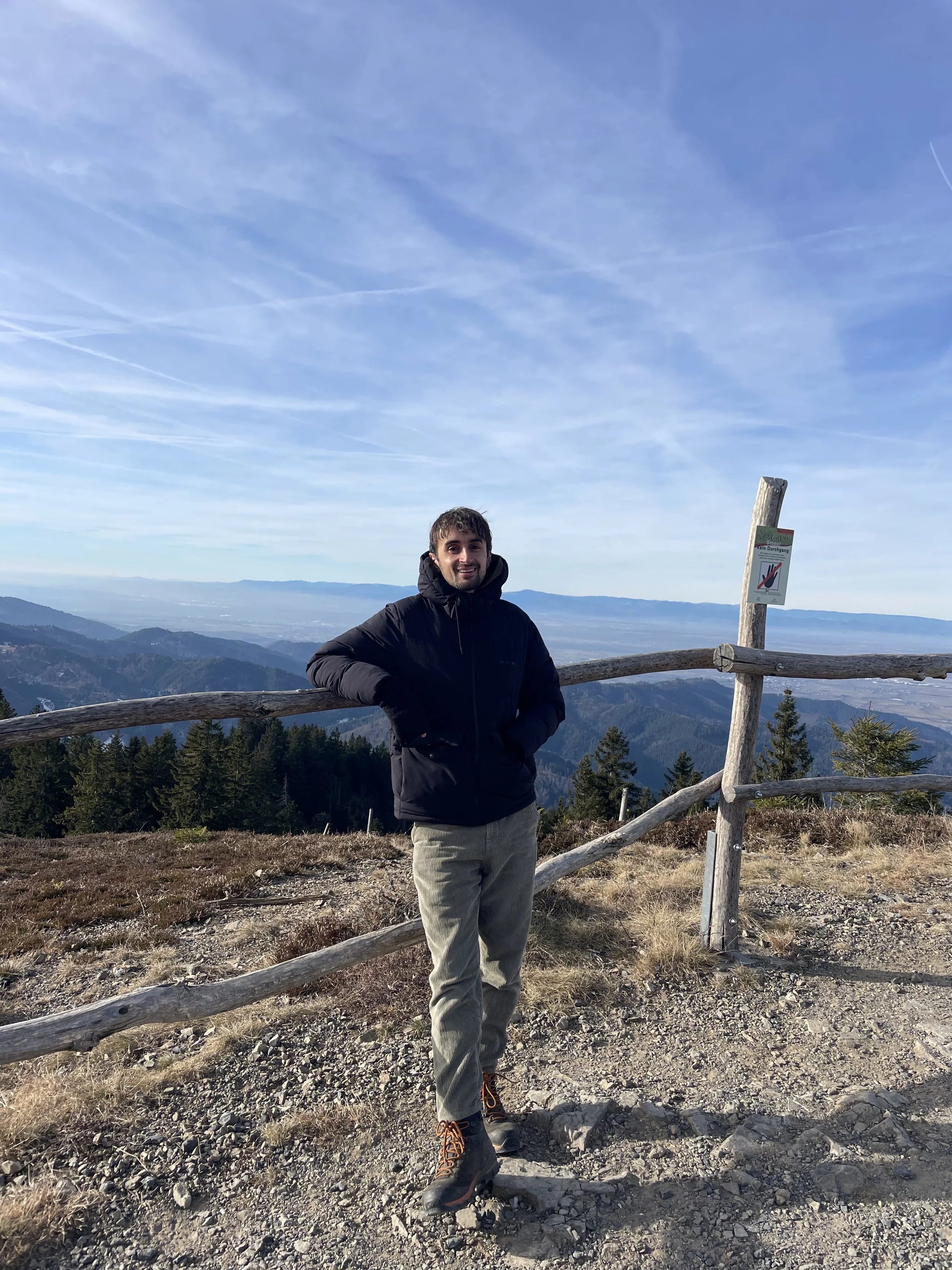 Mann auf einer Bergspitze mit Aussicht auf Berge und Himmel, trägt dunkle Jacke und helle Hosen, steht vor einer Holzzaun.