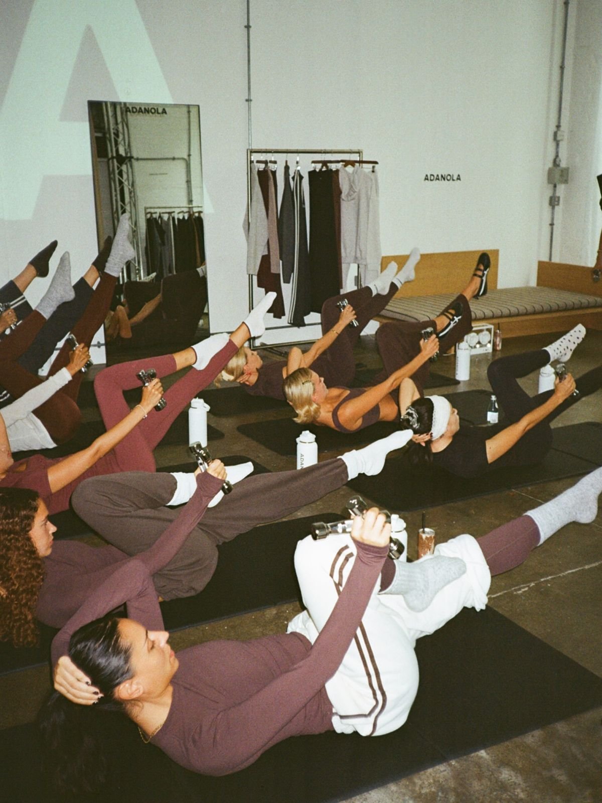 Group of women participating in a fitness or yoga class in a studio, lying on mats, lifting one leg and holding small weights, with water bottles beside them.