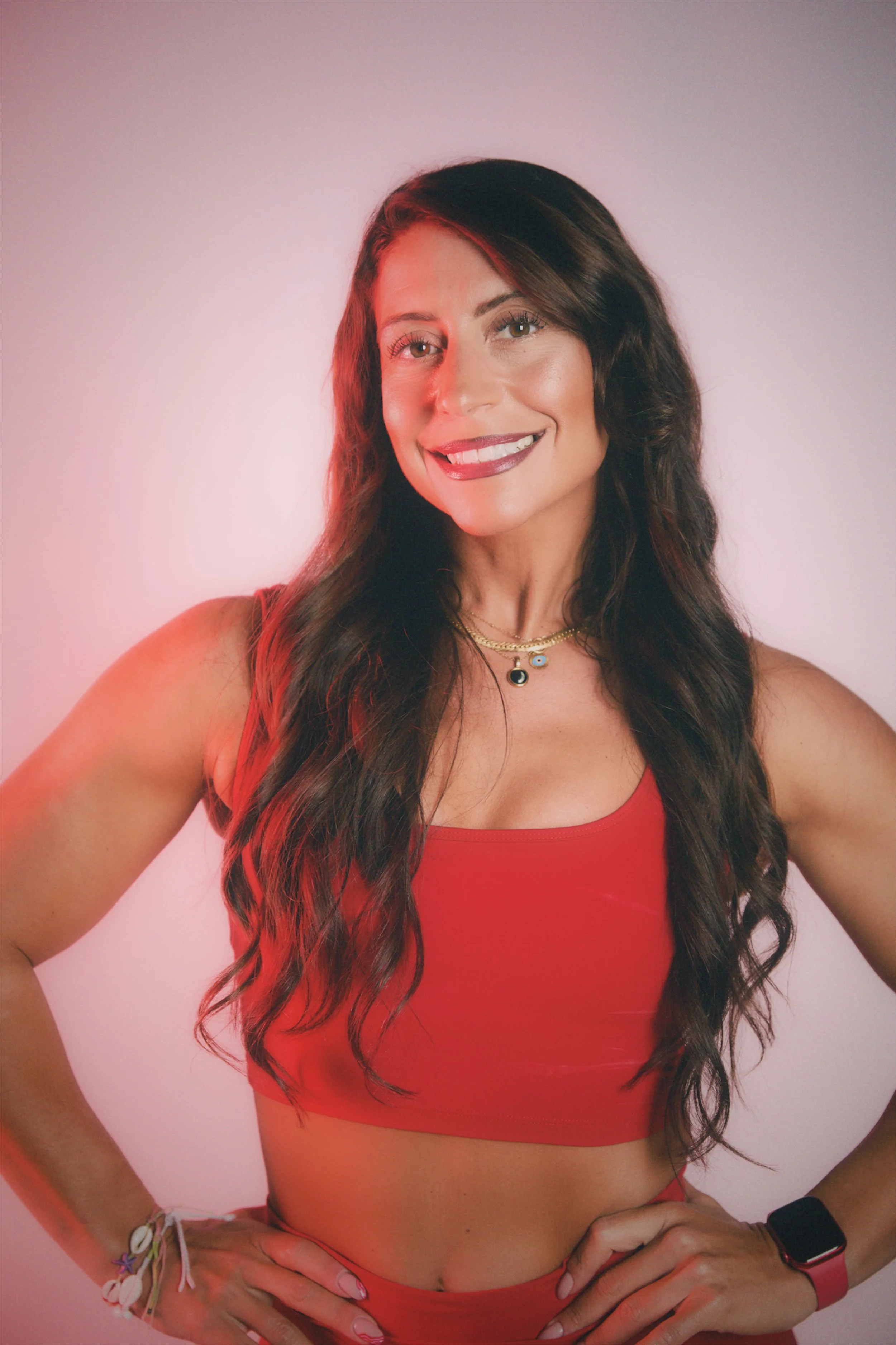 A woman with long wavy brown hair smiling at the camera, wearing a red tank top, multiple bracelets, a necklace, and an Apple Watch, posed with hands on hips.
