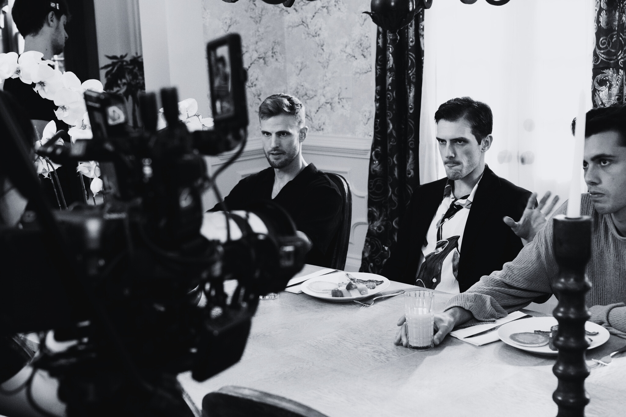 Black and white photo of three men sitting at a dining table, with a camera and crew in the foreground filming the scene, which includes plates of food and drinks on the table, in a well-lit room with curtains and floral wallpaper.