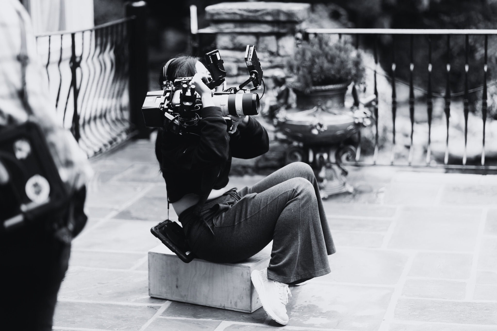 A woman sitting on a small wooden block, operating a professional video camera with two external monitors, outdoors on a patio with a stone floor, a metal railing, and potted plants in the background.