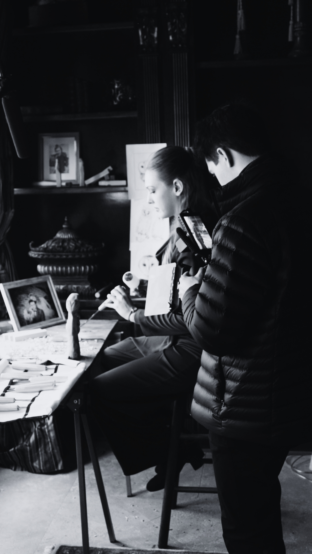 A person is taking a photograph of a woman sitting at a table with various items and framed pictures, with another person standing nearby observing.