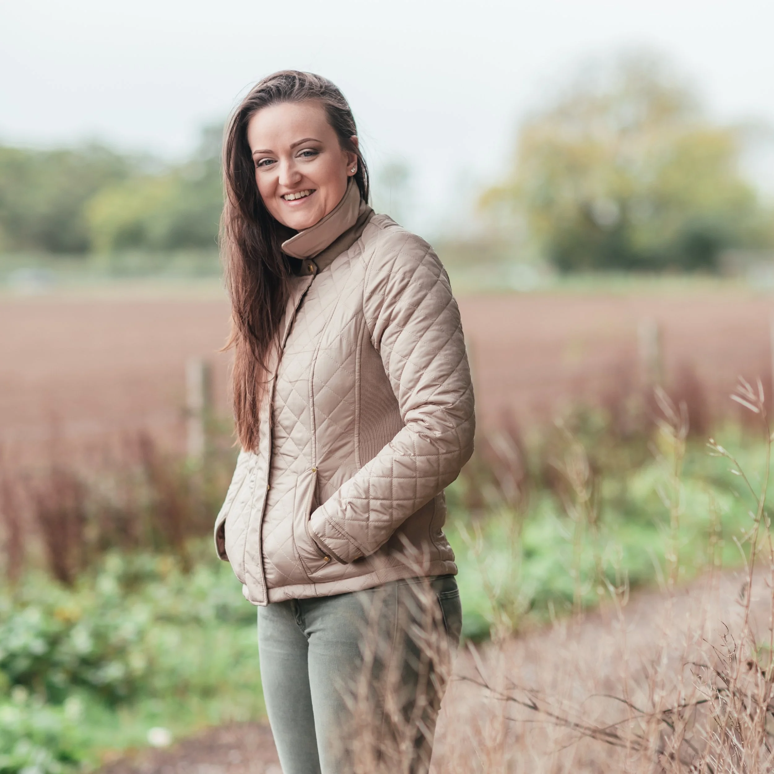 A woman smiling outdoors in a quilted beige jacket with hands in pockets, standing in a field with blurred trees and plants in the background.