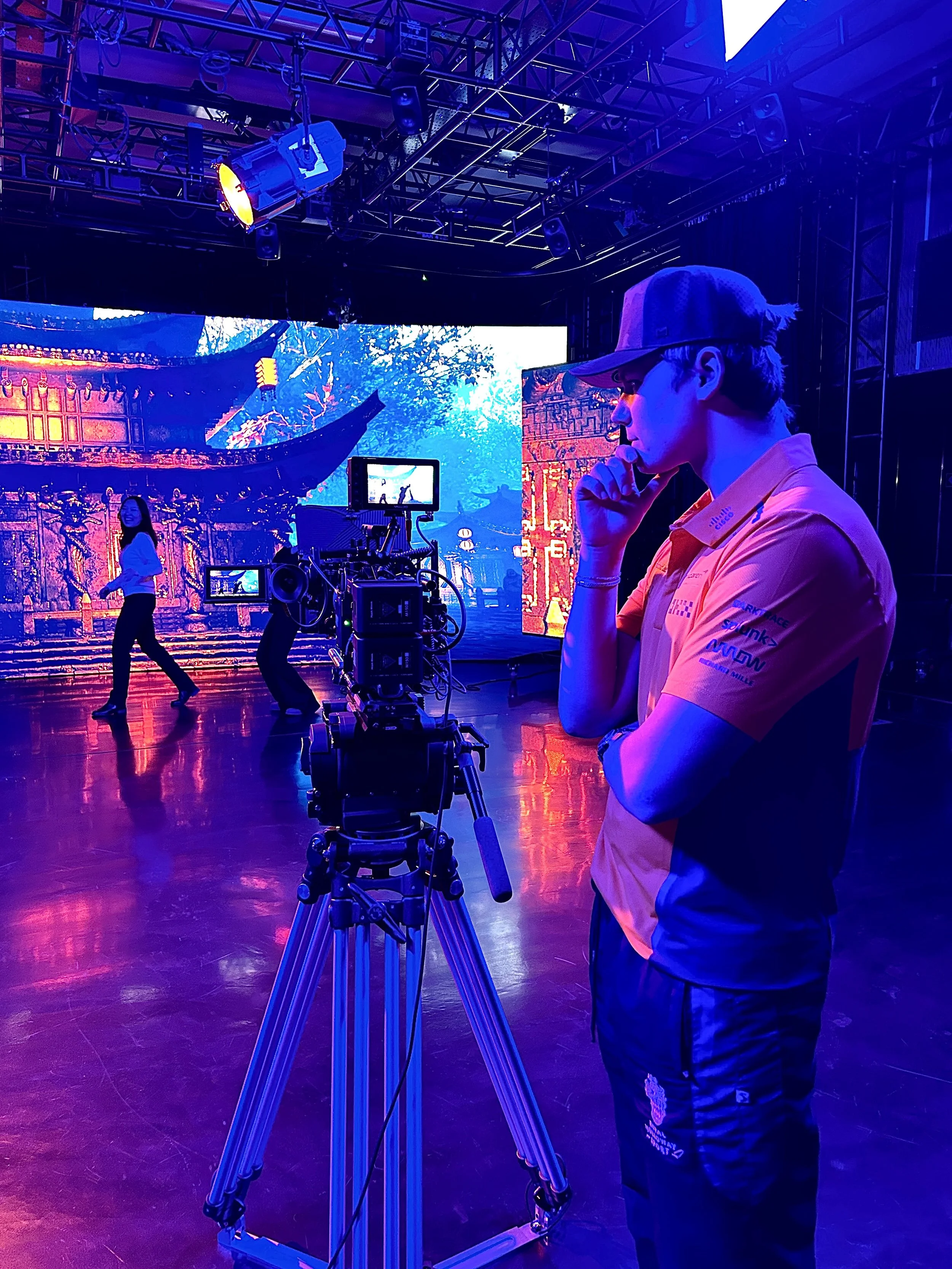 A person in a pink and blue shirt wearing glasses and a cap stands beside a camera on a tripod, observing a stage with dancers and a colorful digital backdrop in a dark studio or theater setting.