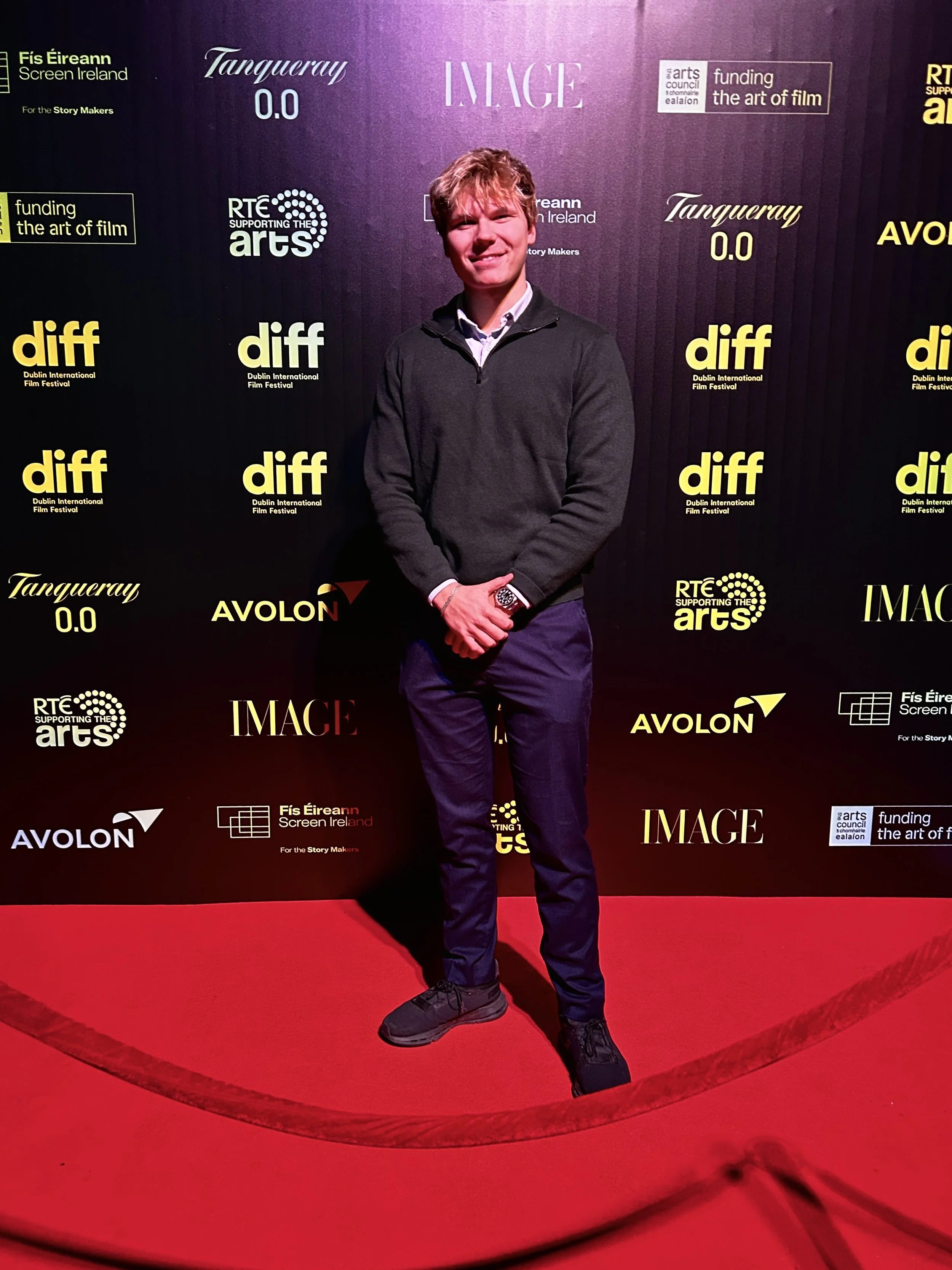 A young man standing on a red carpet at an event, smiling, wearing a dark zip-up jacket, navy pants, and black shoes, with a backdrop featuring logos and text related to the film and arts festival.