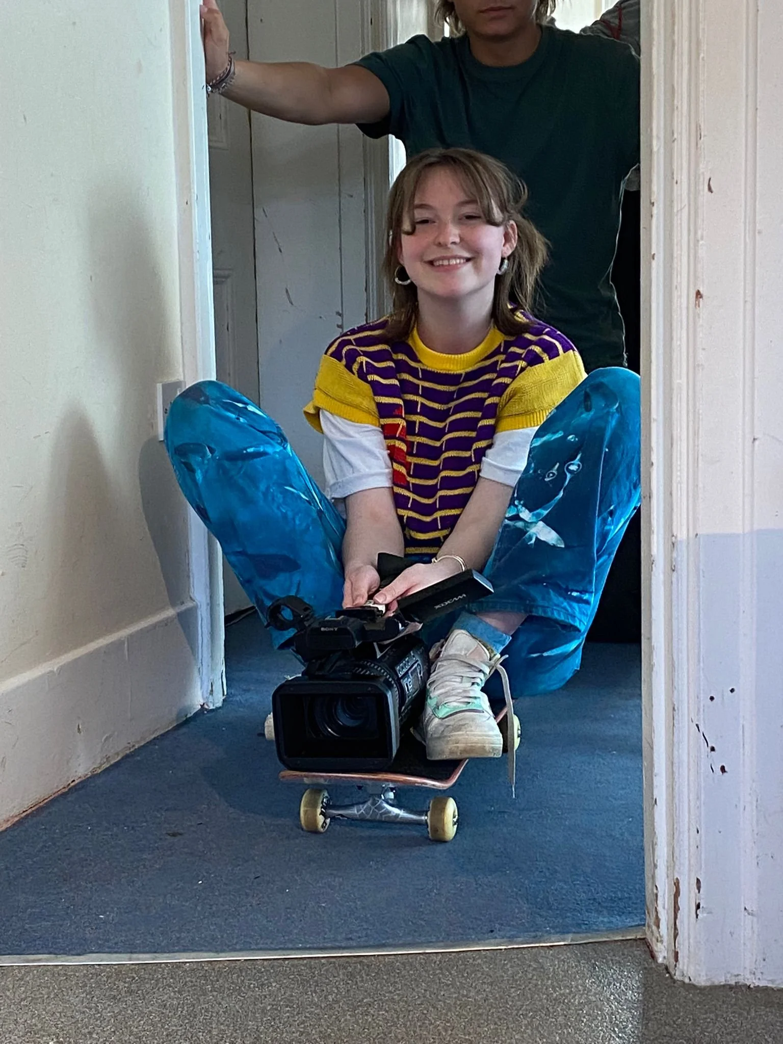A young girl is sitting on a skateboard with a video camera on her lap, inside a doorway, smiling at the camera. An adult is standing behind her, partially visible, holding the doorframe open.