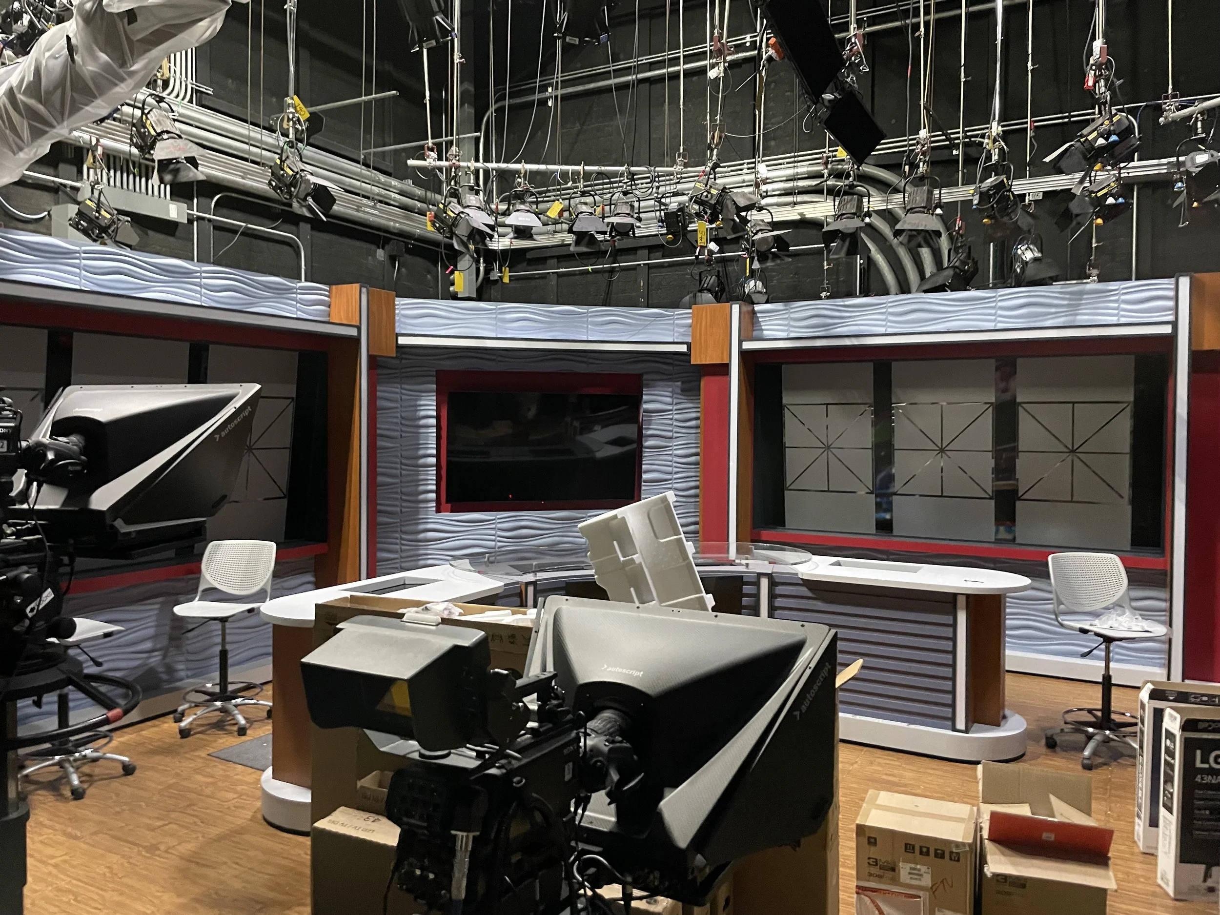 Television news studio with cameras, lighting, chairs, a curved news desk, and a large screen behind the desk.