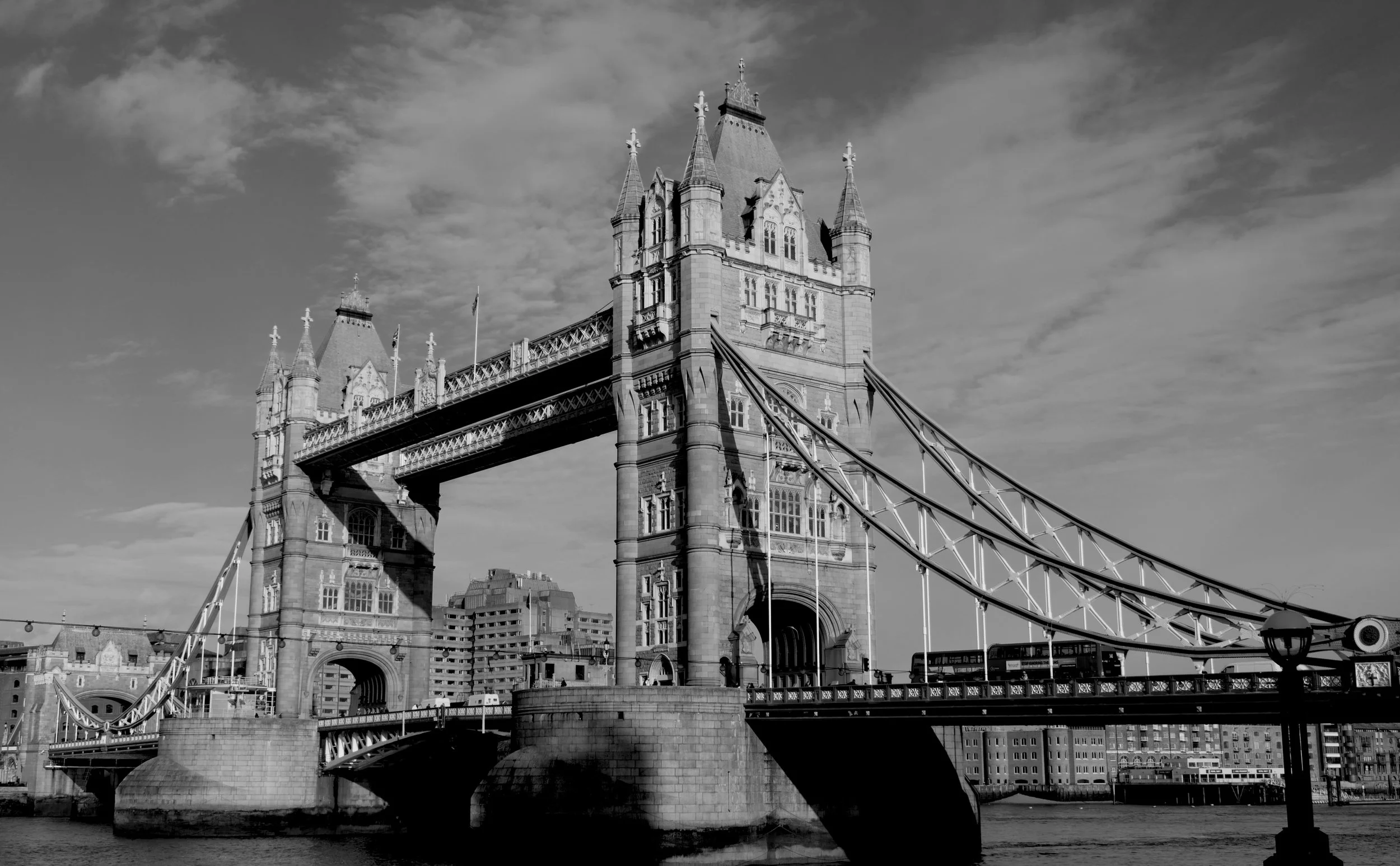 Black and white photo of Tower Bridge in London, showing its two towers connected by walkways and a drawbridge, with some buildings in the background and a partly cloudy sky.
