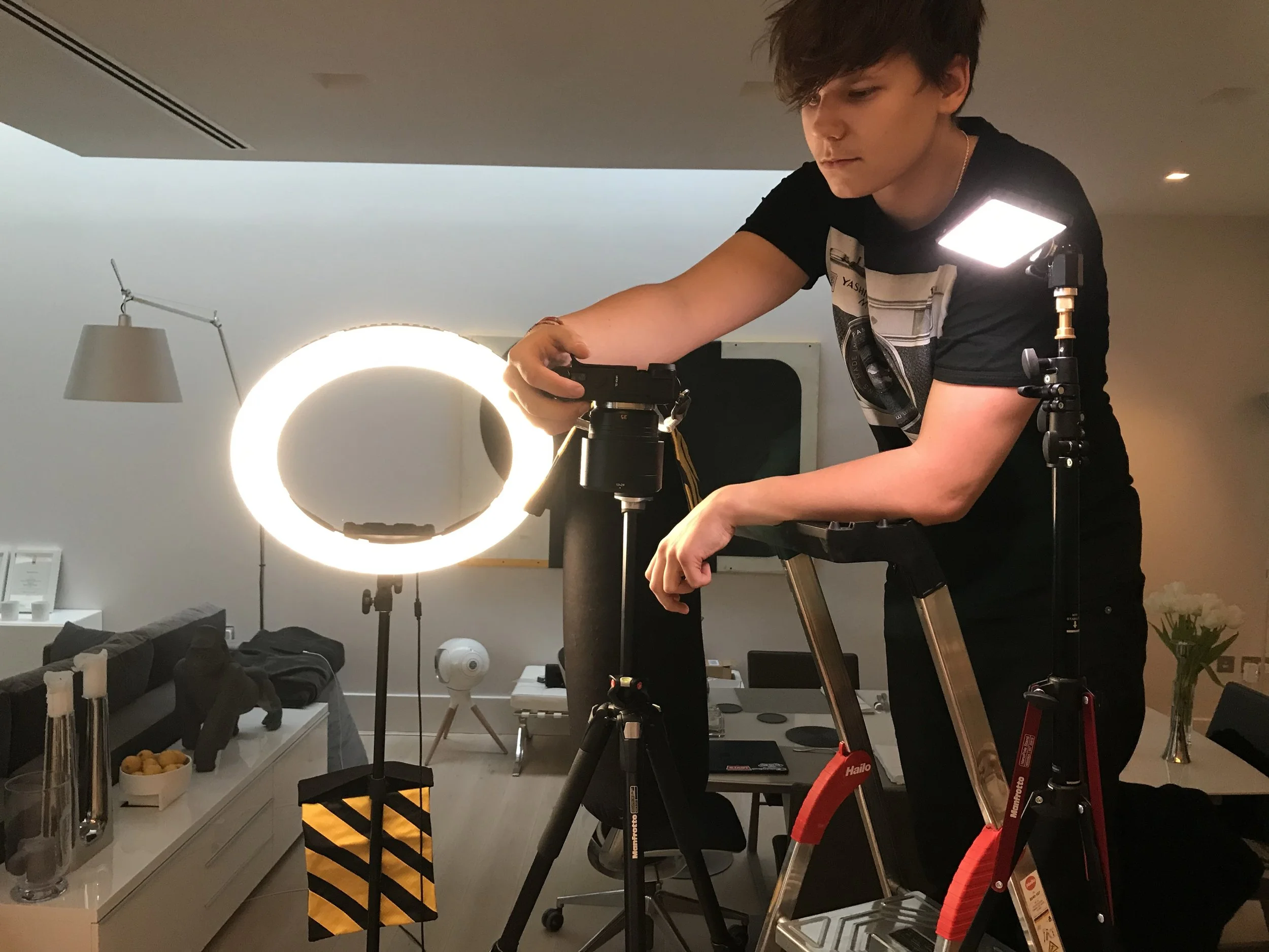 A person standing on a ladder setting up a camera with a ring light illuminating the scene in a room with furniture, a TV, and flowers.
