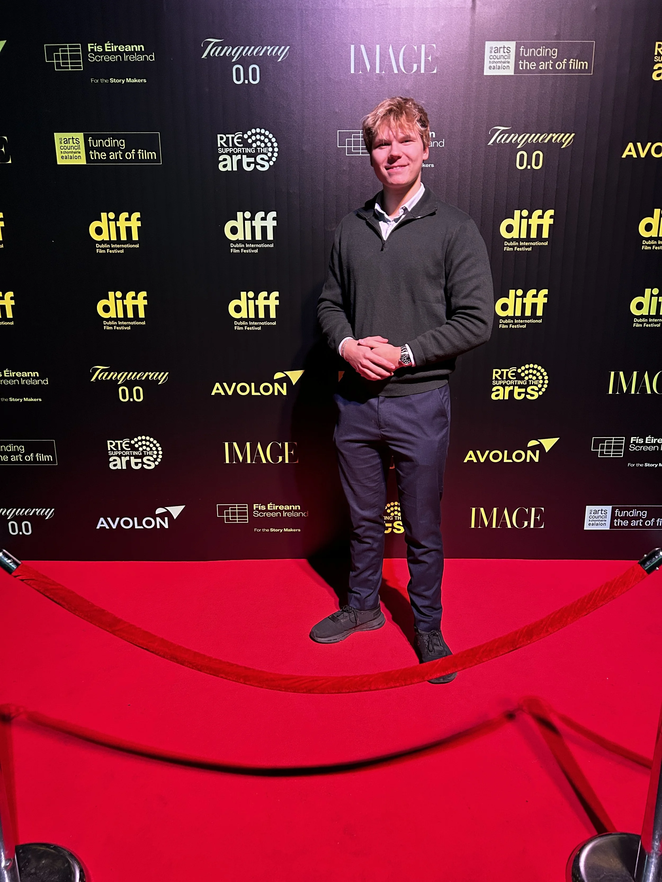 Young man standing on a red carpet at a film event, smiling, with a black backdrop displaying logos of various sponsors and organizations including Dublin International Film Festival, Arts Council, RTÉ, and AVOLON.