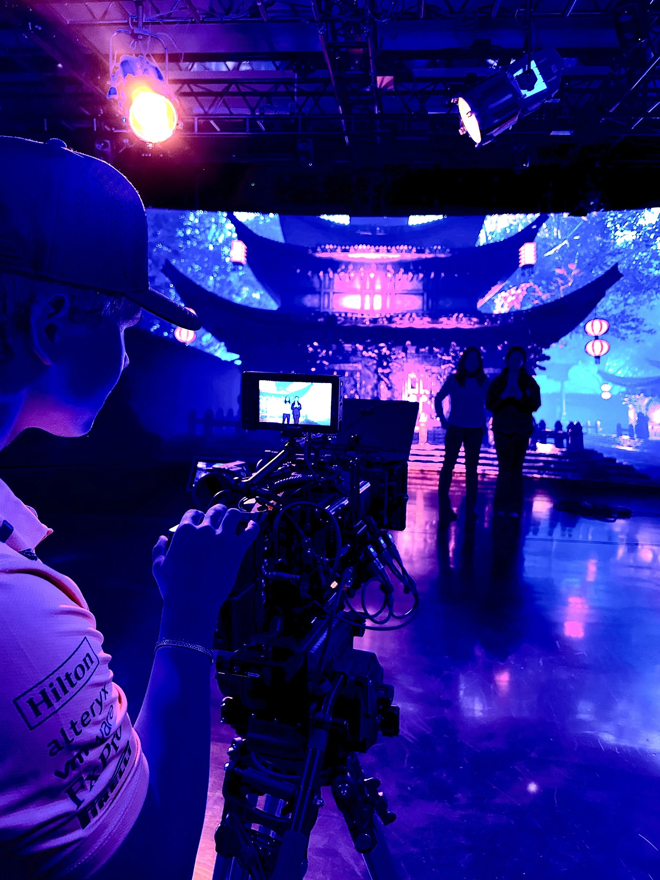 A person operating a film camera on a stage with a blue and purple illuminated backdrop featuring traditional Asian lanterns and architecture, with two people standing and talking in silhouette in the distance.