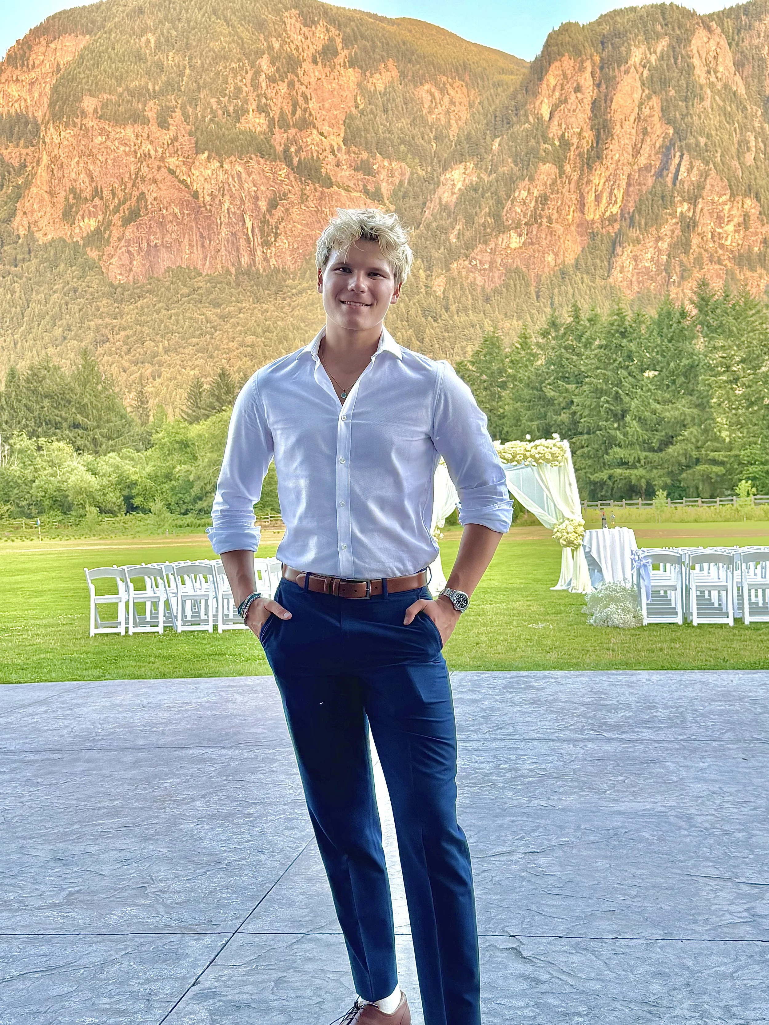 Young man in white dress shirt and blue pants standing outdoors at what appears to be a wedding venue with mountainous landscape, green trees, and white chairs and decorations in the background.