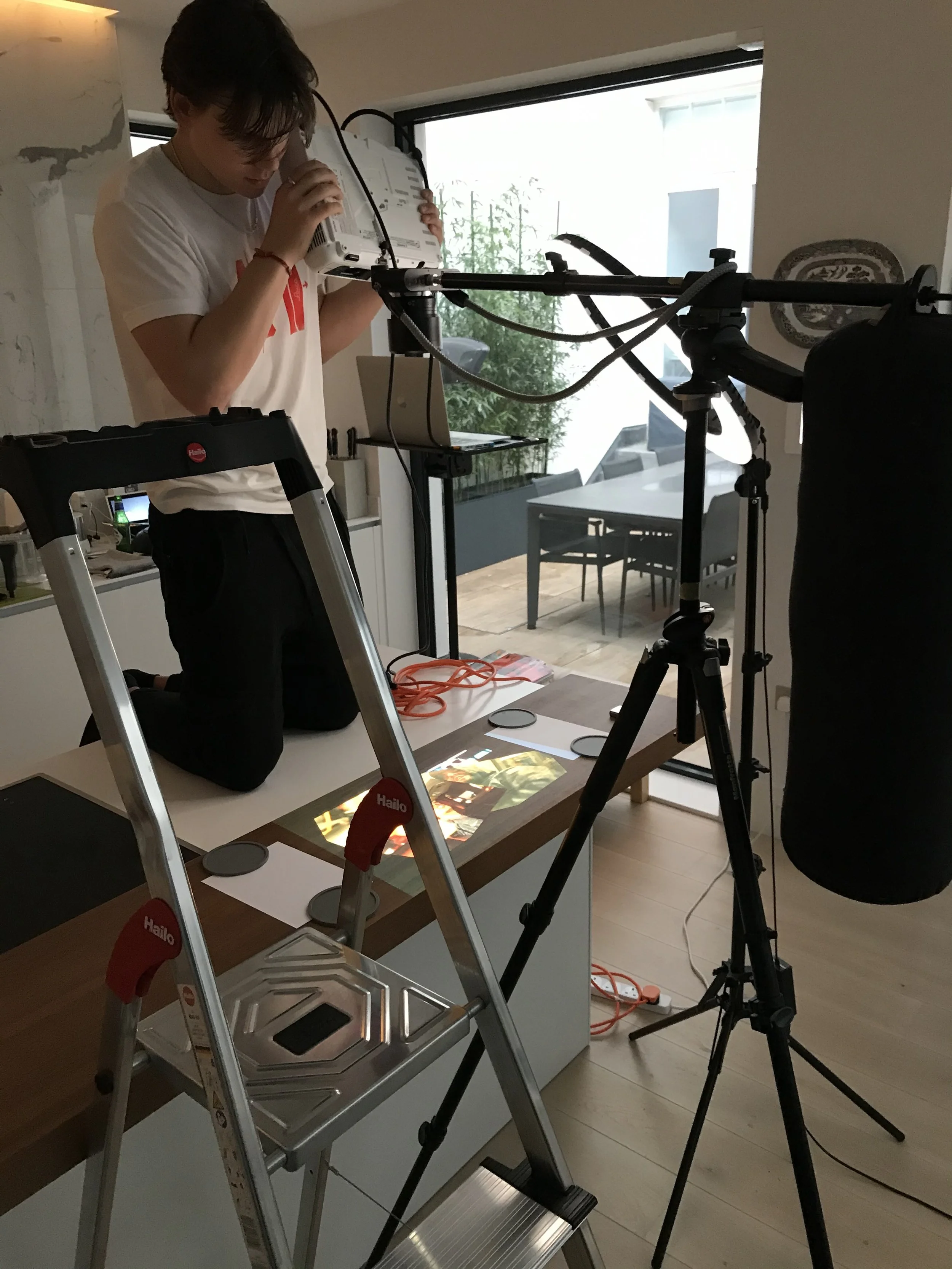 A person kneeling on a chair, adjusting a camera or projector mounted on a tripod, with a large screen or display on a table showing a colorful image, in a room with large windows and outdoor patio furniture.