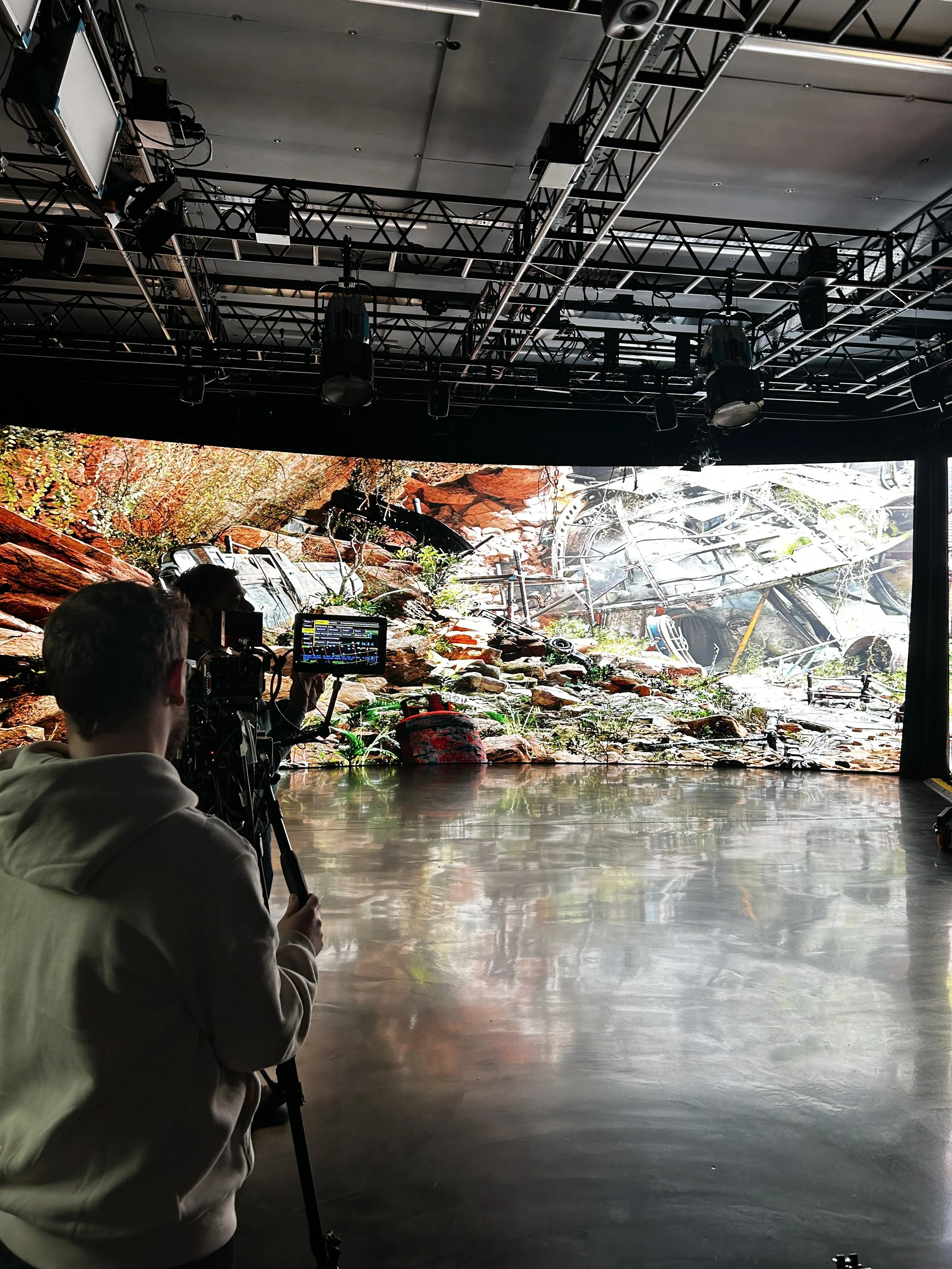 A behind-the-scenes view of a creation set with a large digital backdrop displaying a rugged landscape with rocks, logs, and a crashed airplane. A person operates a camera on a tripod, filming or photographing the scene.
