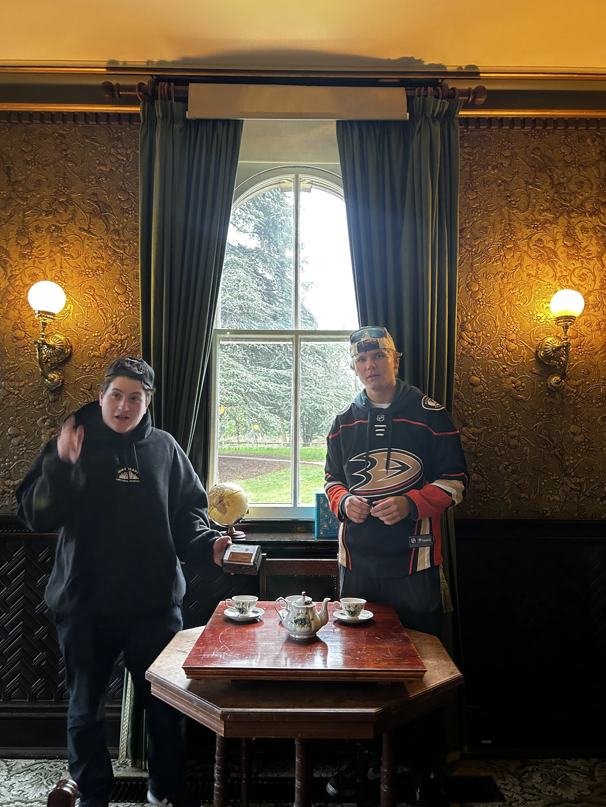 Two young boys standing in a vintage room with ornate wallpaper, green curtains, and wall sconces. They are positioned near a window with outdoor greenery visible. One boy is holding a globe and a small box, the other is wearing a hockey jersey and a cap. A teapot and two cups are on a small wooden table in front of them.