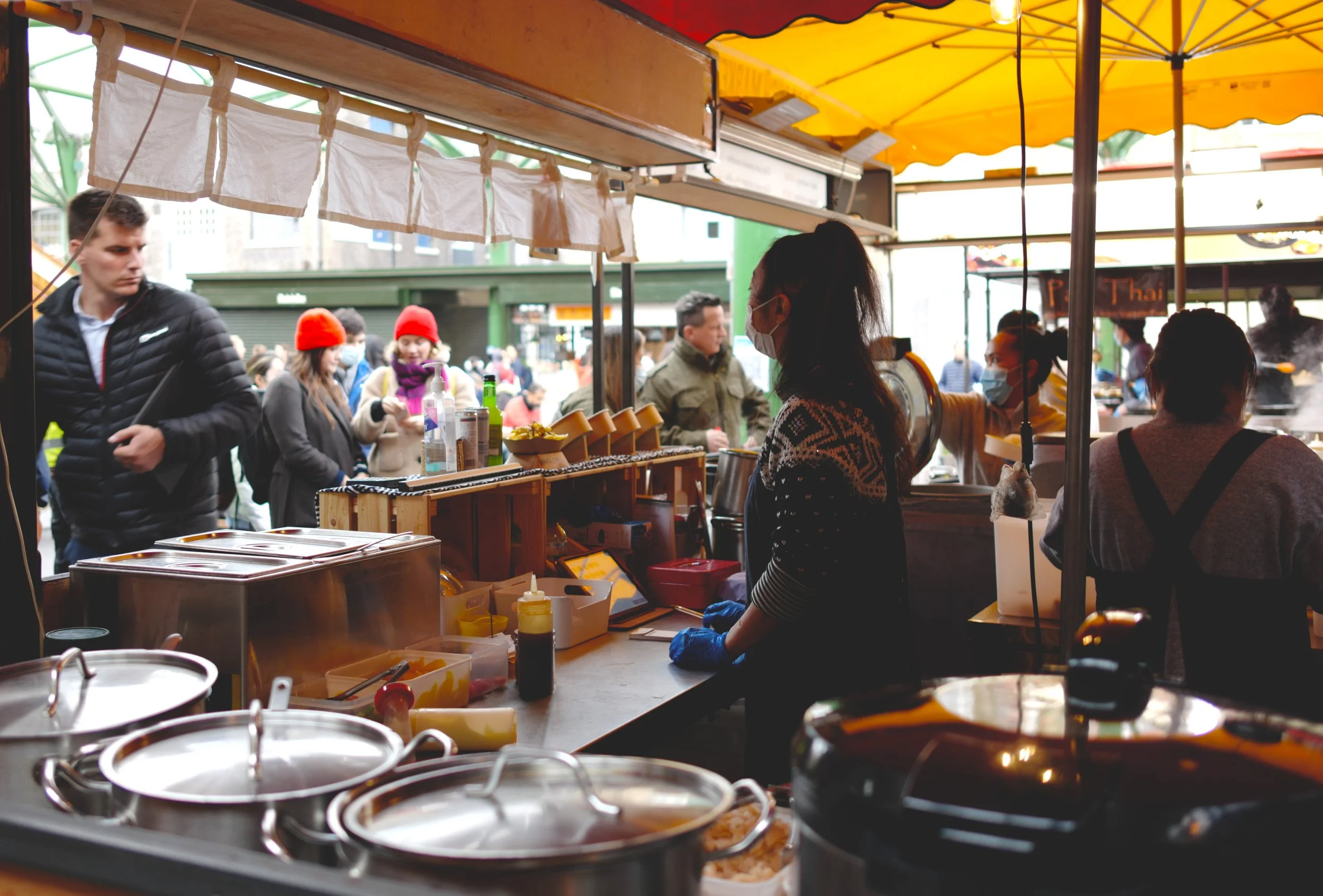 Food stall at a street market with vendors and customers, some wearing masks, under a yellow canopy.