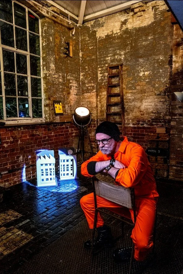 A man wearing an orange jumpsuit, black beanie, and glasses sitting on a chair in a rustic, brick-walled room with an industrial style, with a ladder leaning against the wall, and two small illuminated art pieces resembling buildings on the floor.