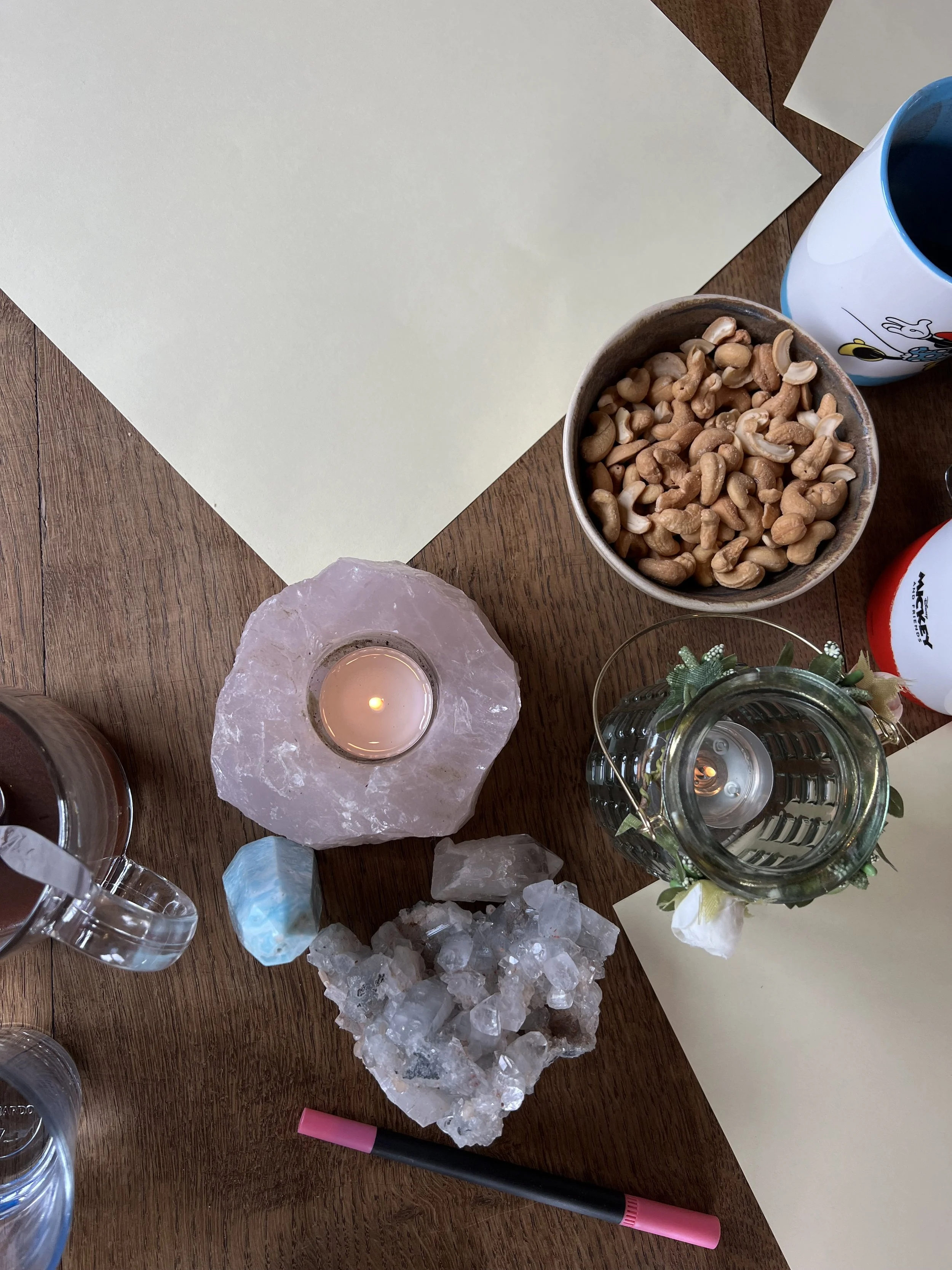 Een tafel met een vaas, een kaars op een rozenkwarts stand, een kom cashewnoten, een glas, een pen, twee foliecups en enkele losse kristallen.