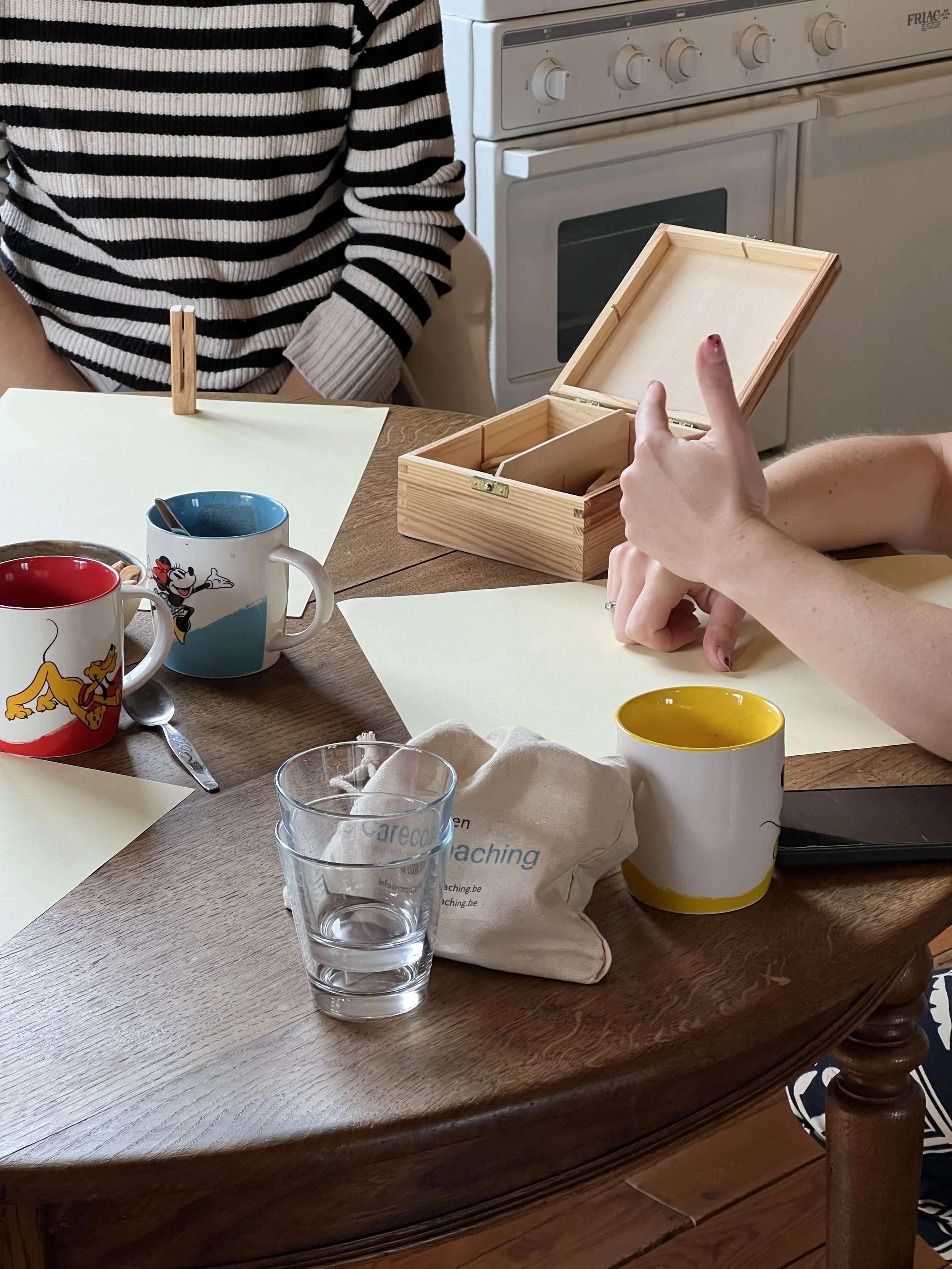 Een houten tafel met meerdere lege tekenpapier vellen, drie koffie- of theemokken, een glas water, een wit doekje en een smartphone. Er zitten ook mensen aan de tafel. In de achtergrond een oven en keukenkastjes.