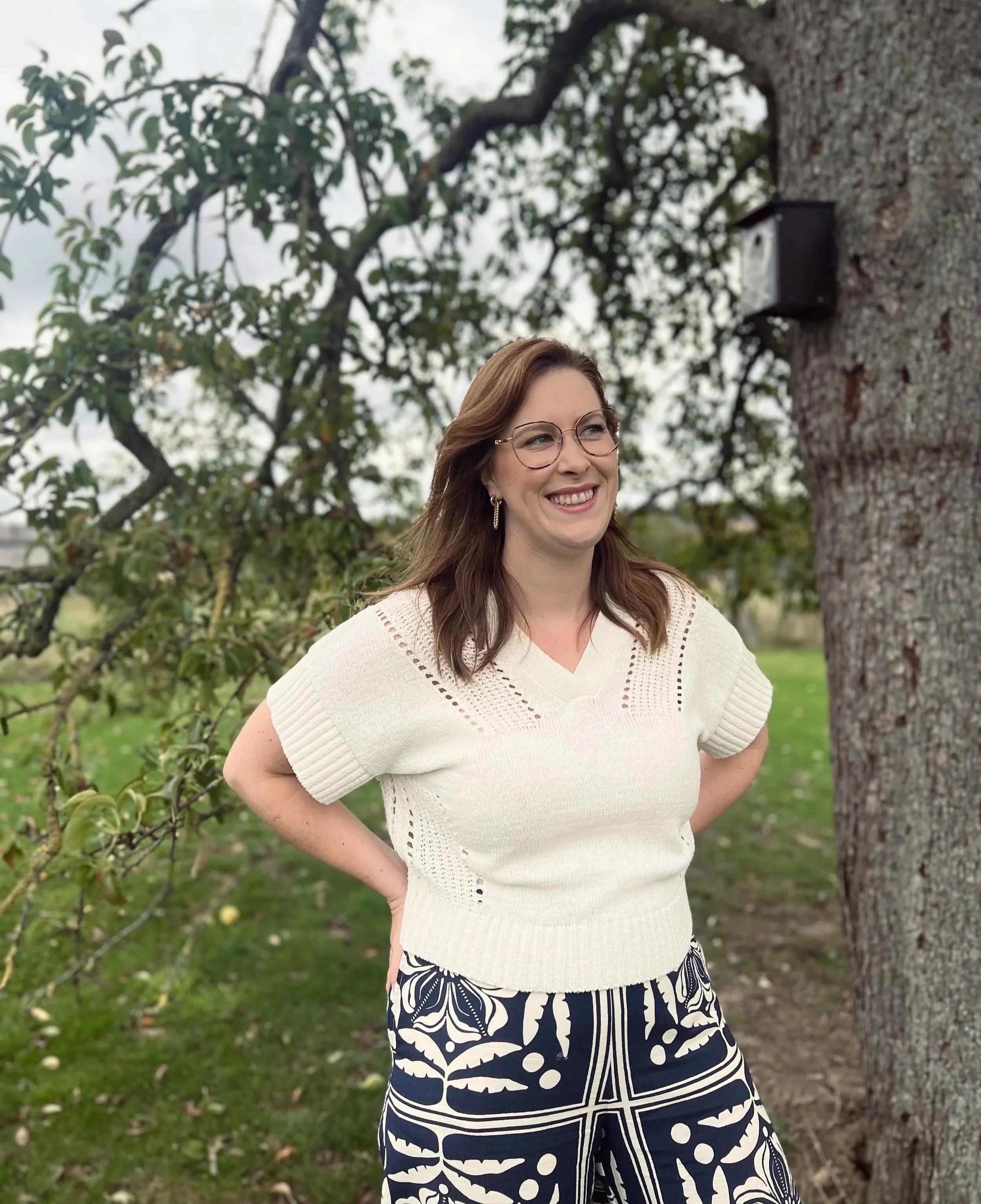 Vrouw met bruin haar, bril en witte trui die glimlacht en buiten staat bij een boom, in een groene tuin.