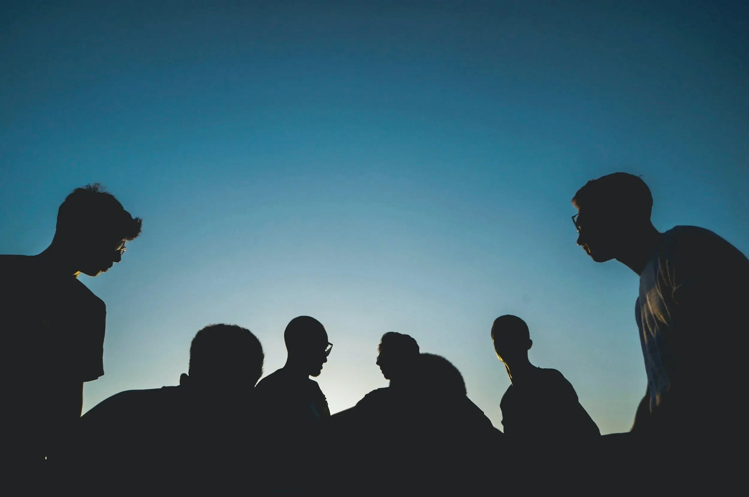 Silhouetten van zes mensen die in groepsdiscussie zitten tegen een blauwe hemel tijdens zonsondergang.