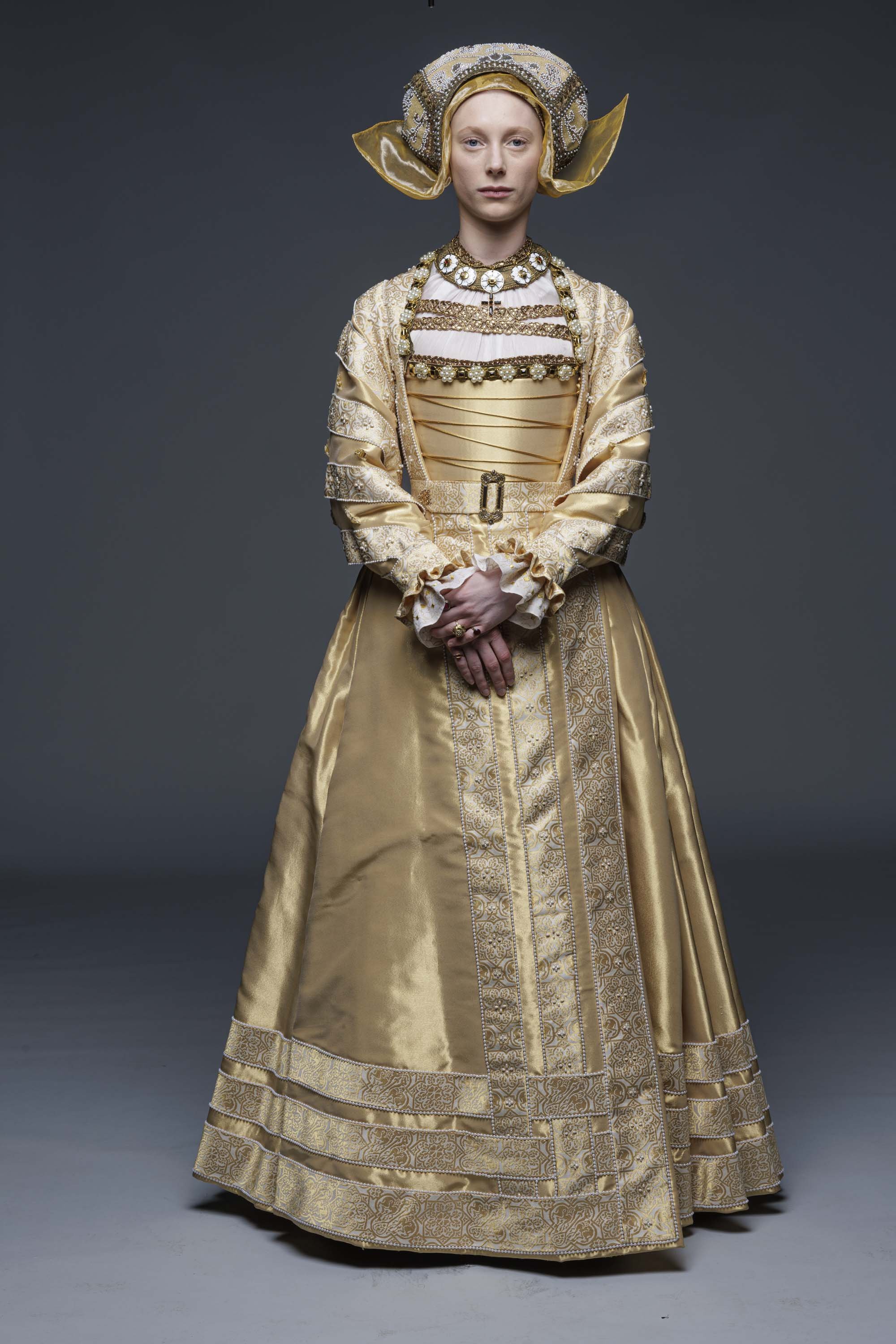 A woman dressed in an elaborate gold historical costume with intricate embroidery, lace, and jewelry, wearing a large ornate headdress.