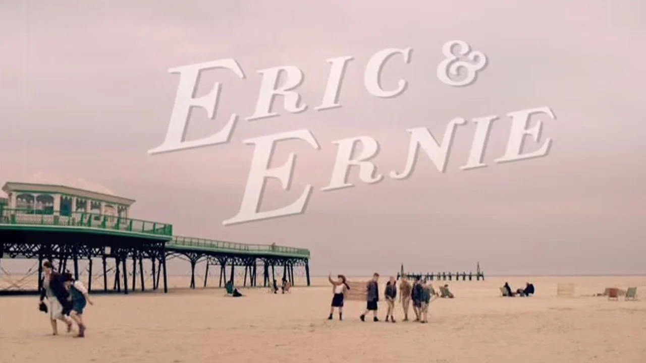 A beach scene with a pier, people walking and sitting on the sand, and the words 'Eric & Ernie' in large white letters across the sky.