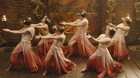 Six women dressed in matching pink and peach dresses and floral headbands perform a synchronized dance with arms extended, in a rustic, warmly lit setting.