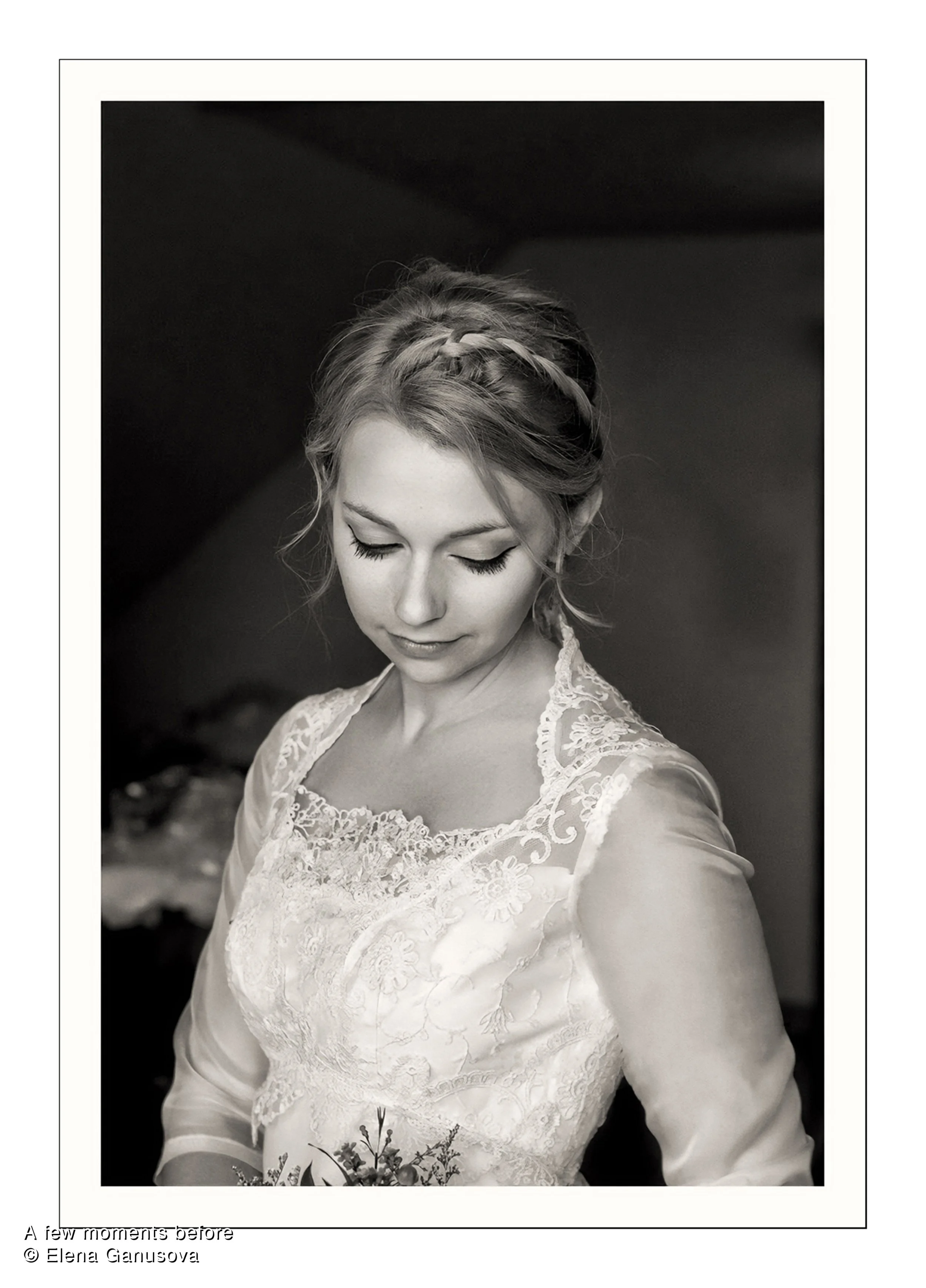 Black and white photograph of a young woman with braided hair, wearing a lace wedding dress, looking down with a serene expression.