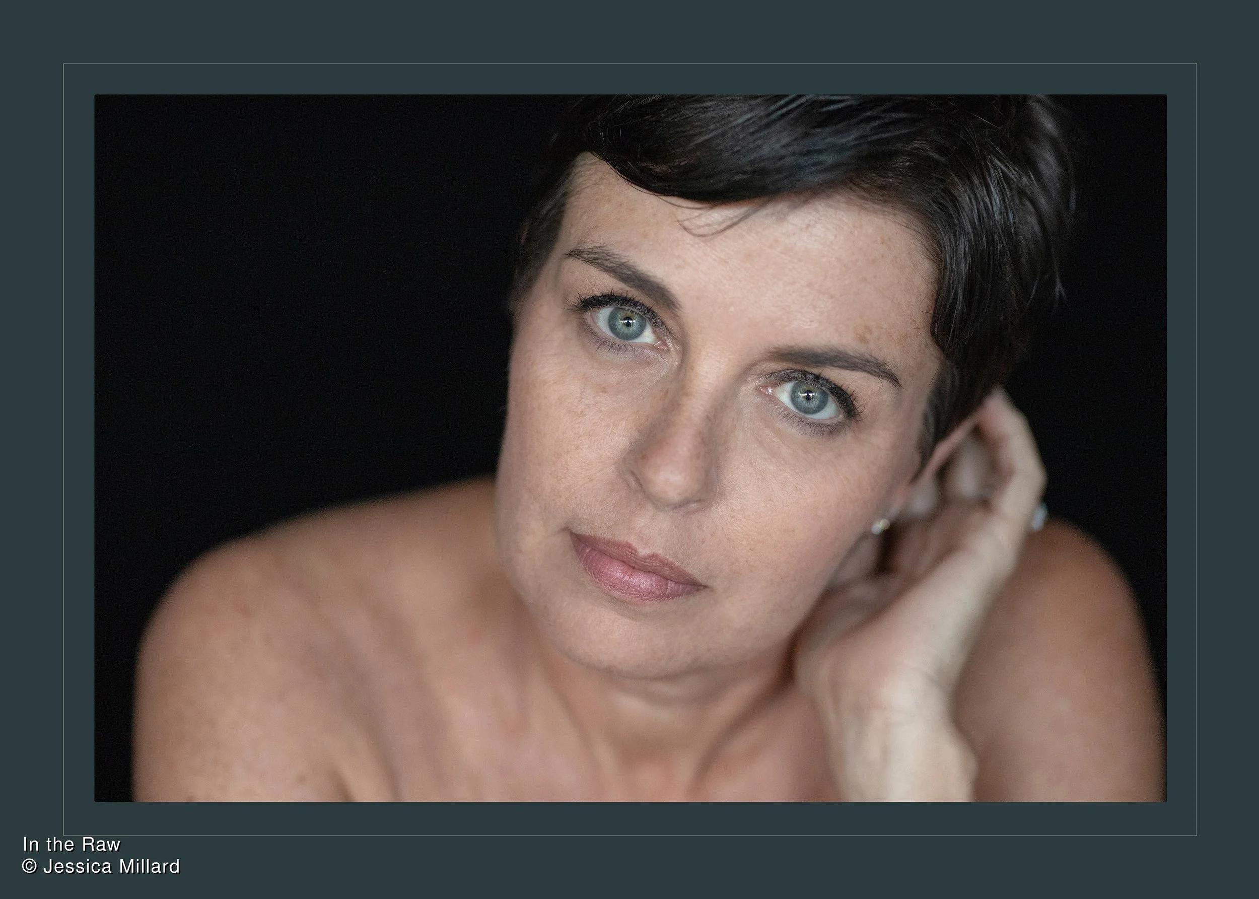 Close-up portrait of a woman with short dark hair and blue eyes, touching her ear against a black background.