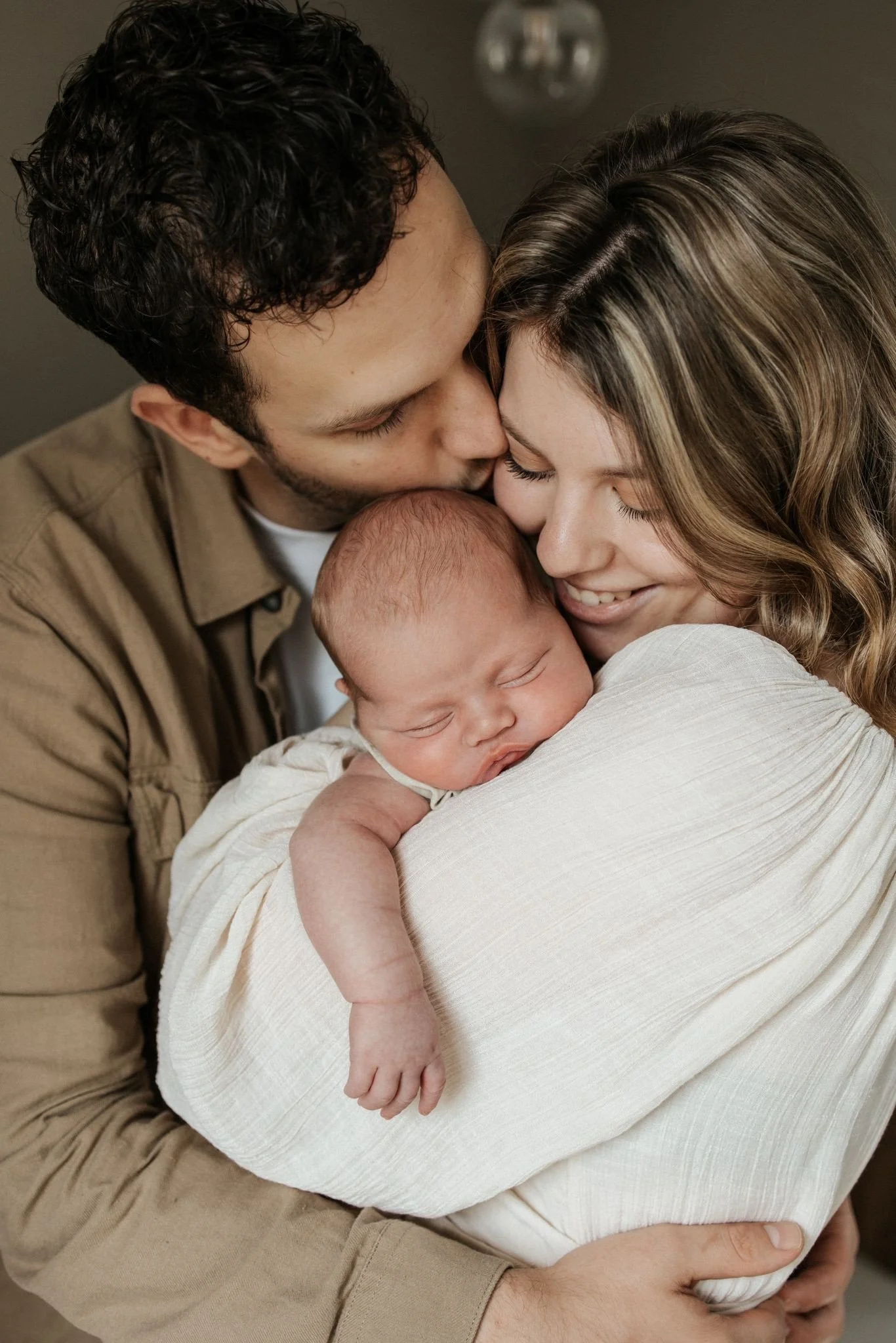 A family of three: a man, a woman, and a newborn baby, all smiling and embracing each other warmly